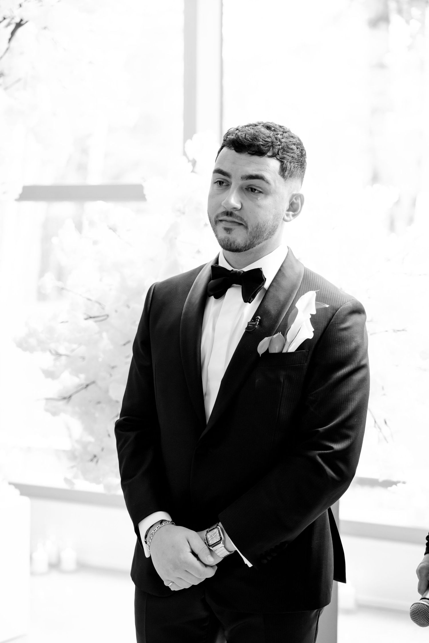 A groom in a classic black tuxedo and bow tie stands waiting during a wedding ceremony.