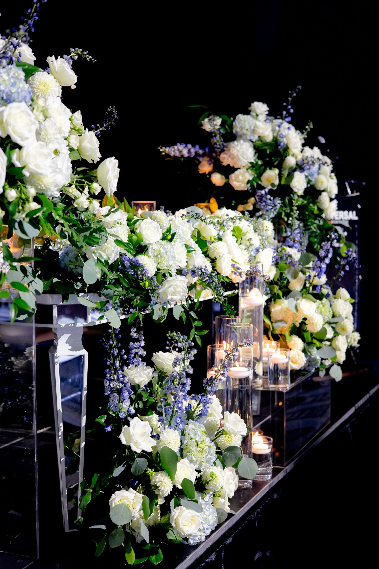 Lavish floral arrangement with white roses and purple accents on mirrored display stand.