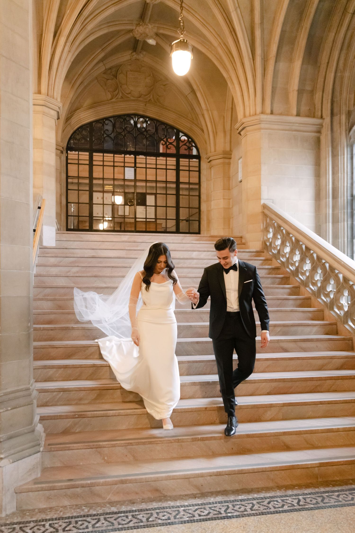 A couple descends an ornate stone staircase in formal wedding attire with vaulted ceilings above.