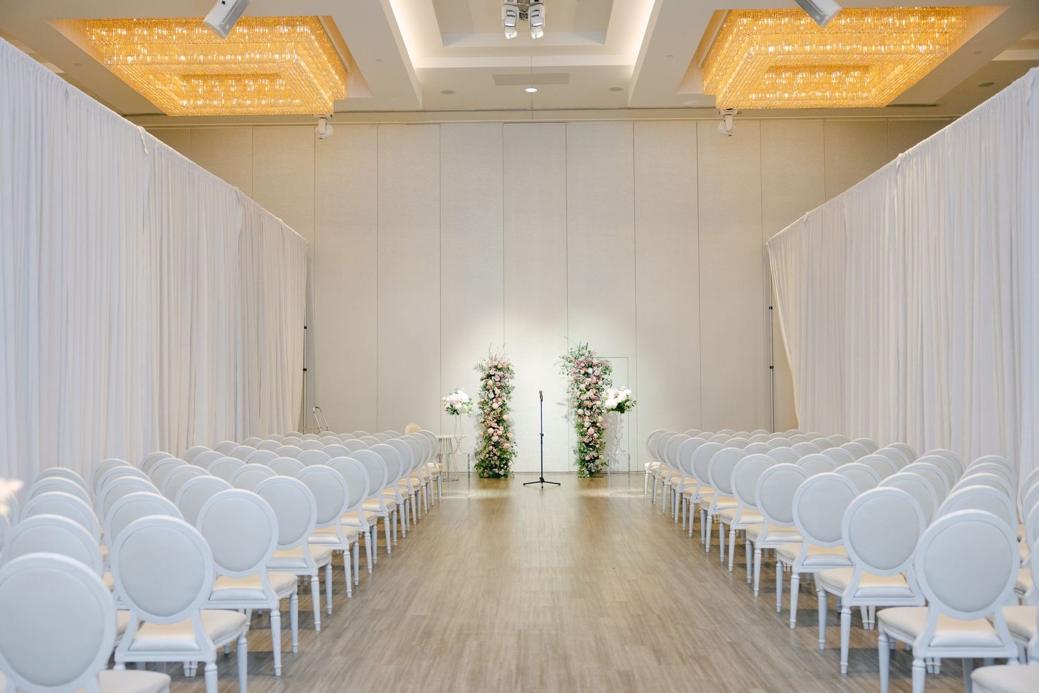 Modern white wedding ceremony space with elegant chairs and warm overhead lighting.