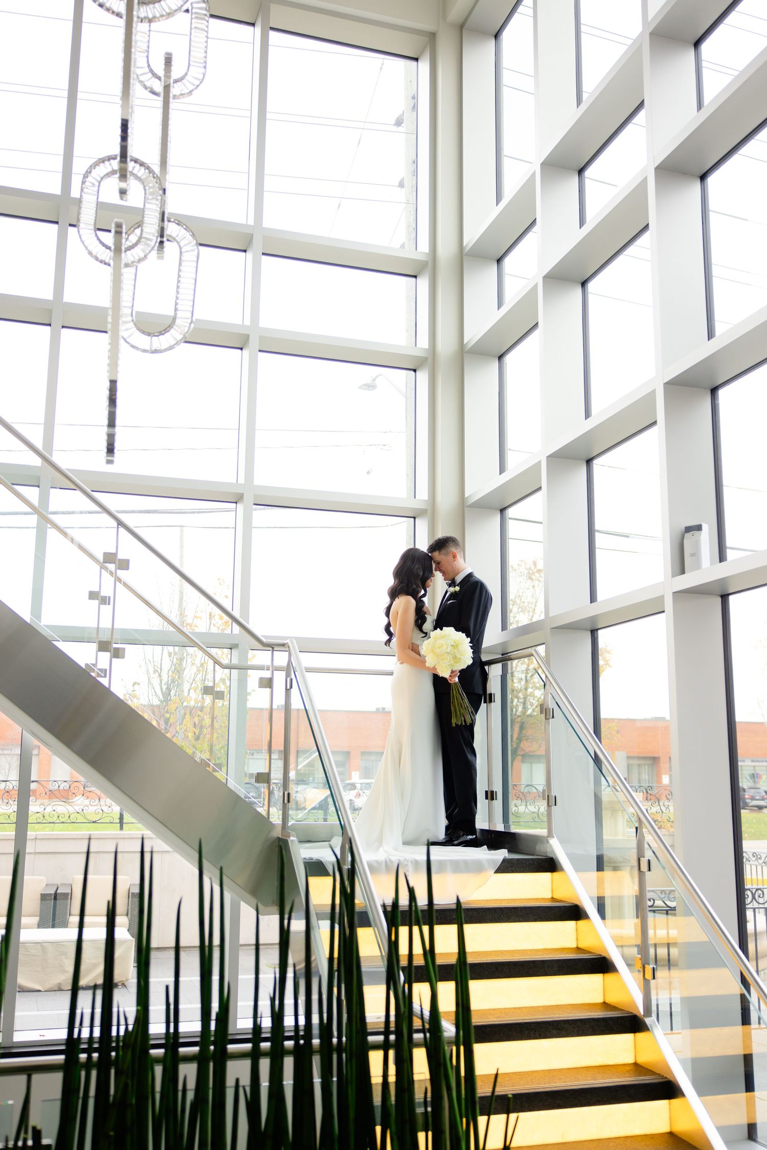 A wedding couple embraces on modern yellow stairs with dramatic floor-to-ceiling windows and hanging light fixtures.