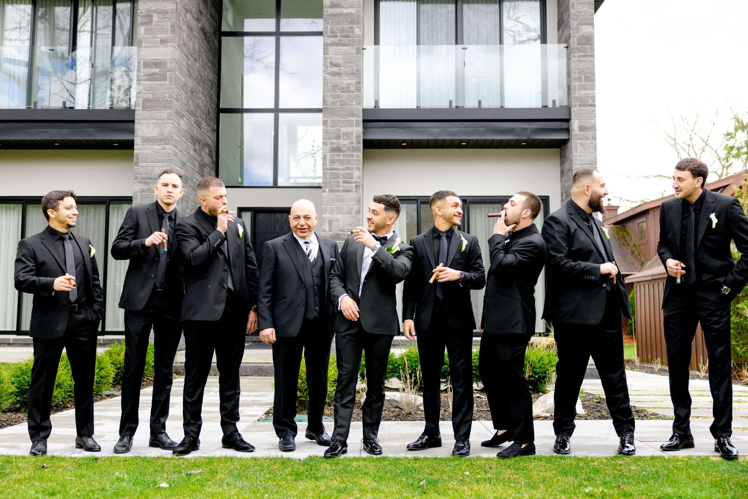 Wedding party in black suits posing casually on grass outside a modern building.