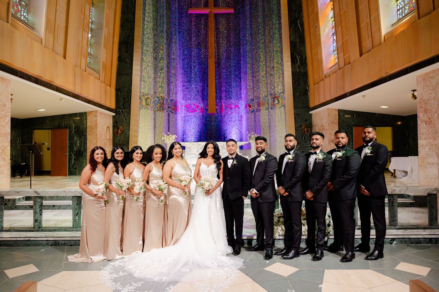 Large wedding party lines up in front of church altar with purple lighting and cross.