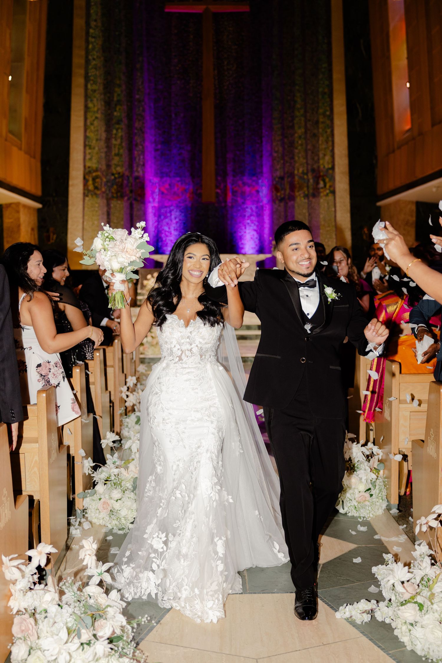 A joyful couple exits their wedding ceremony down a church aisle decorated with white flowers.