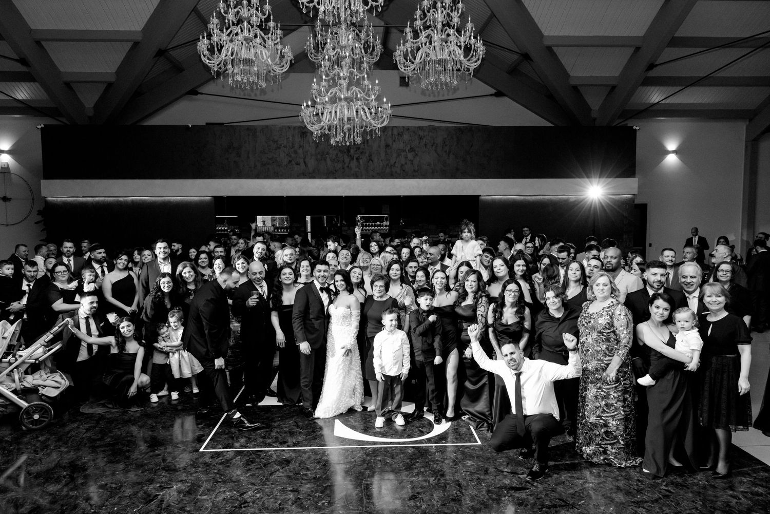 Large wedding celebration group photo with crystal chandeliers overhead in black and white.