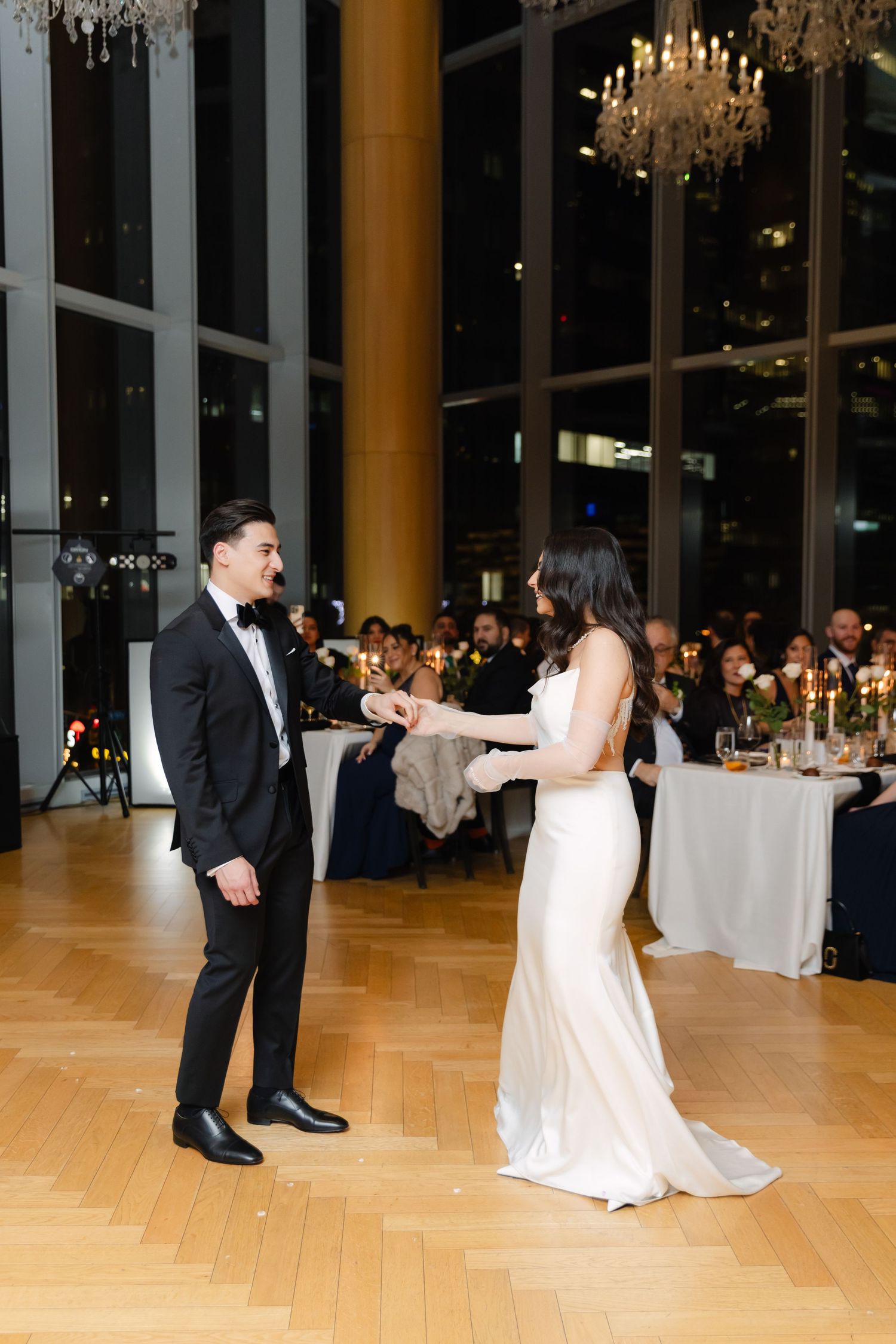Couple shares their first dance in elegant ballroom with crystal chandeliers and city views.