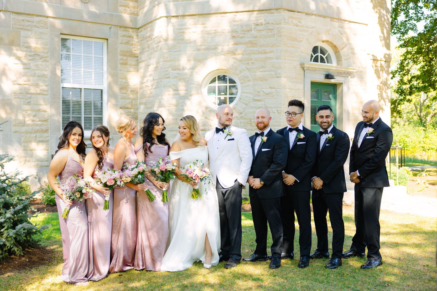 Bridal party in pink gowns and black suits pose for formal photo outside historic stone building.