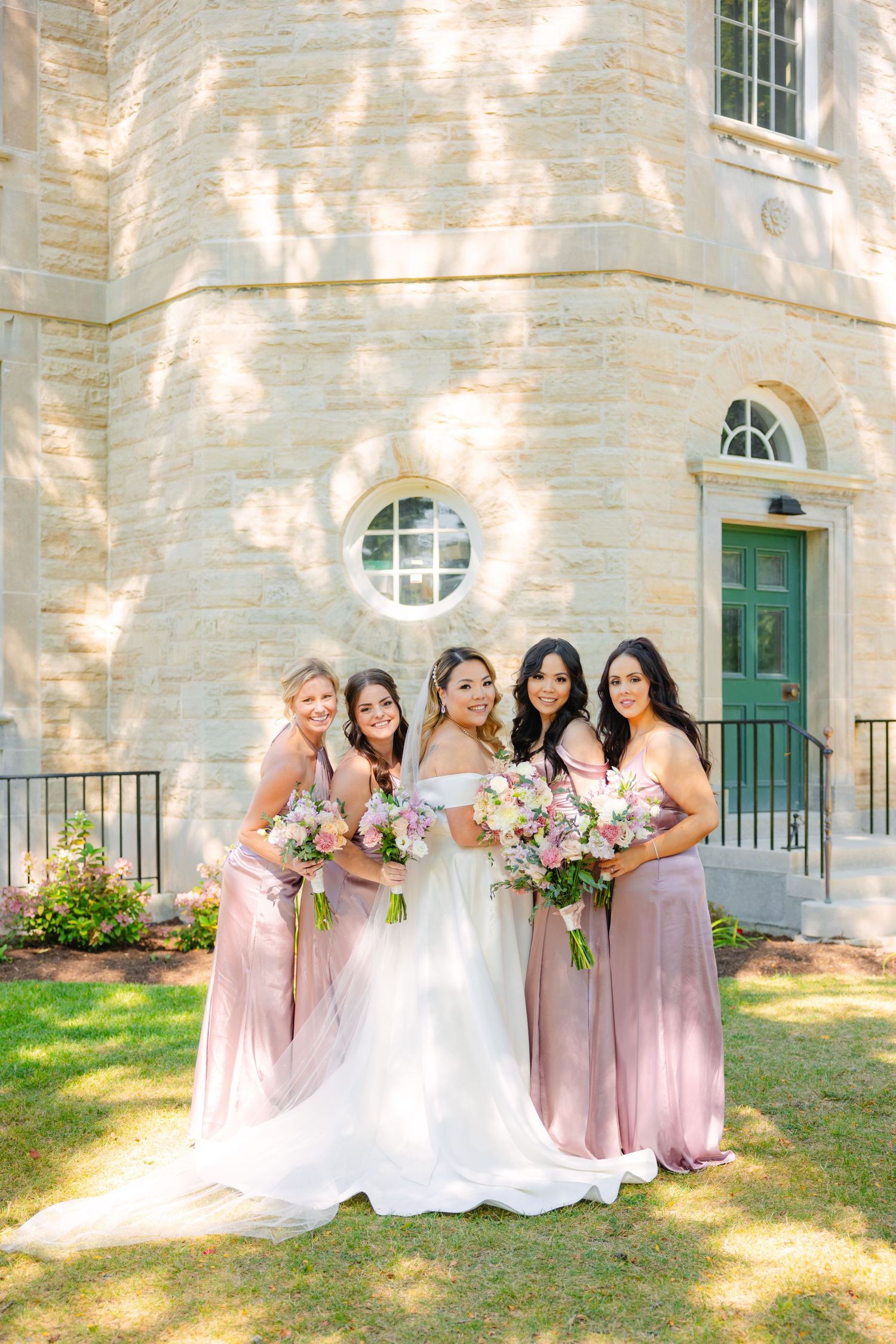 Bridal party in soft pink dresses poses together on lawn in front of stone building.