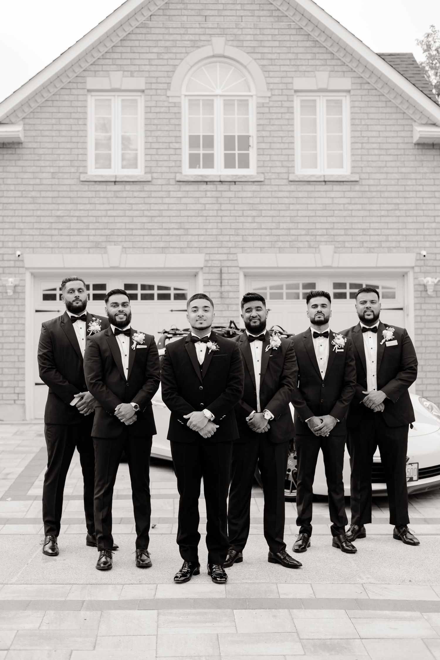 Groomsmen in black tuxedos and bow ties pose formally in front of a beige house with arched windows.