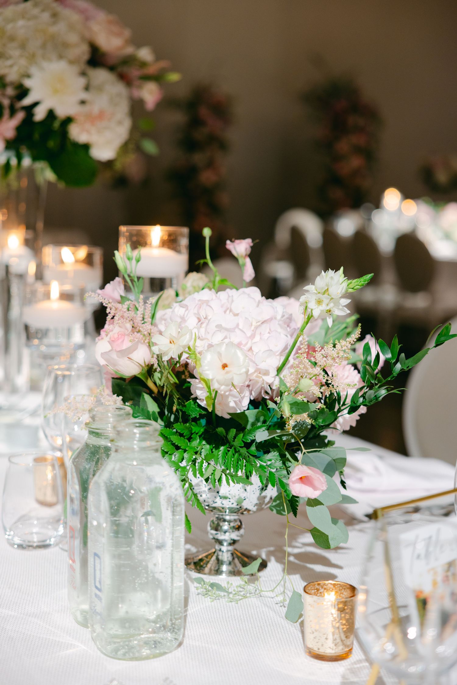 Romantic table decor featuring blush pink flowers, glass bottles, and glowing candles on white tablecloth.