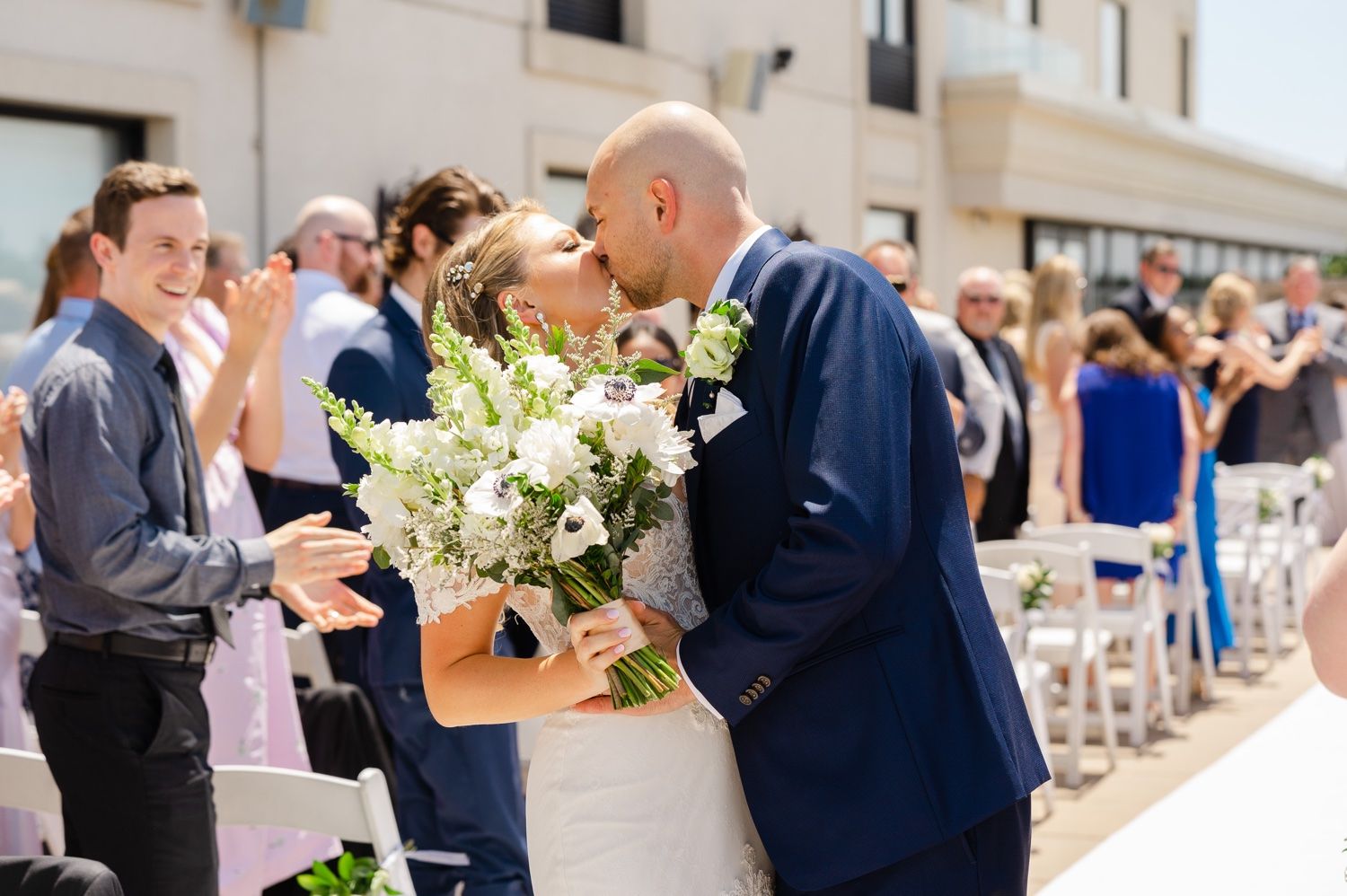 Wedding couple shares intimate moment during outdoor ceremony surrounded by guests and white floral arrangements.