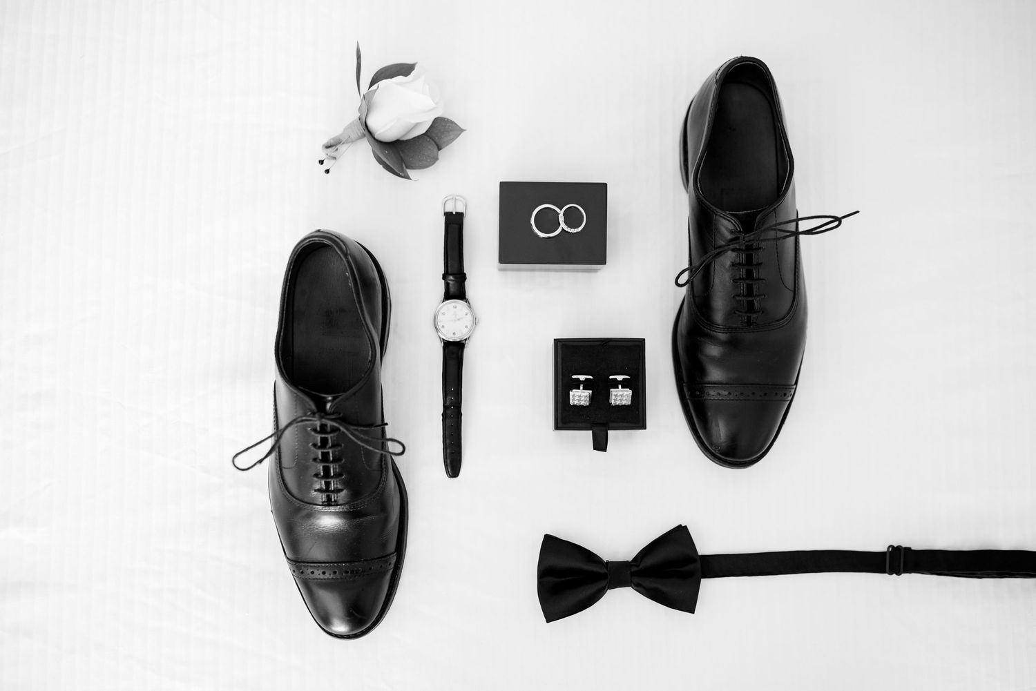 Classic groom accessories including black dress shoes, bow tie, and wedding rings in monochrome.