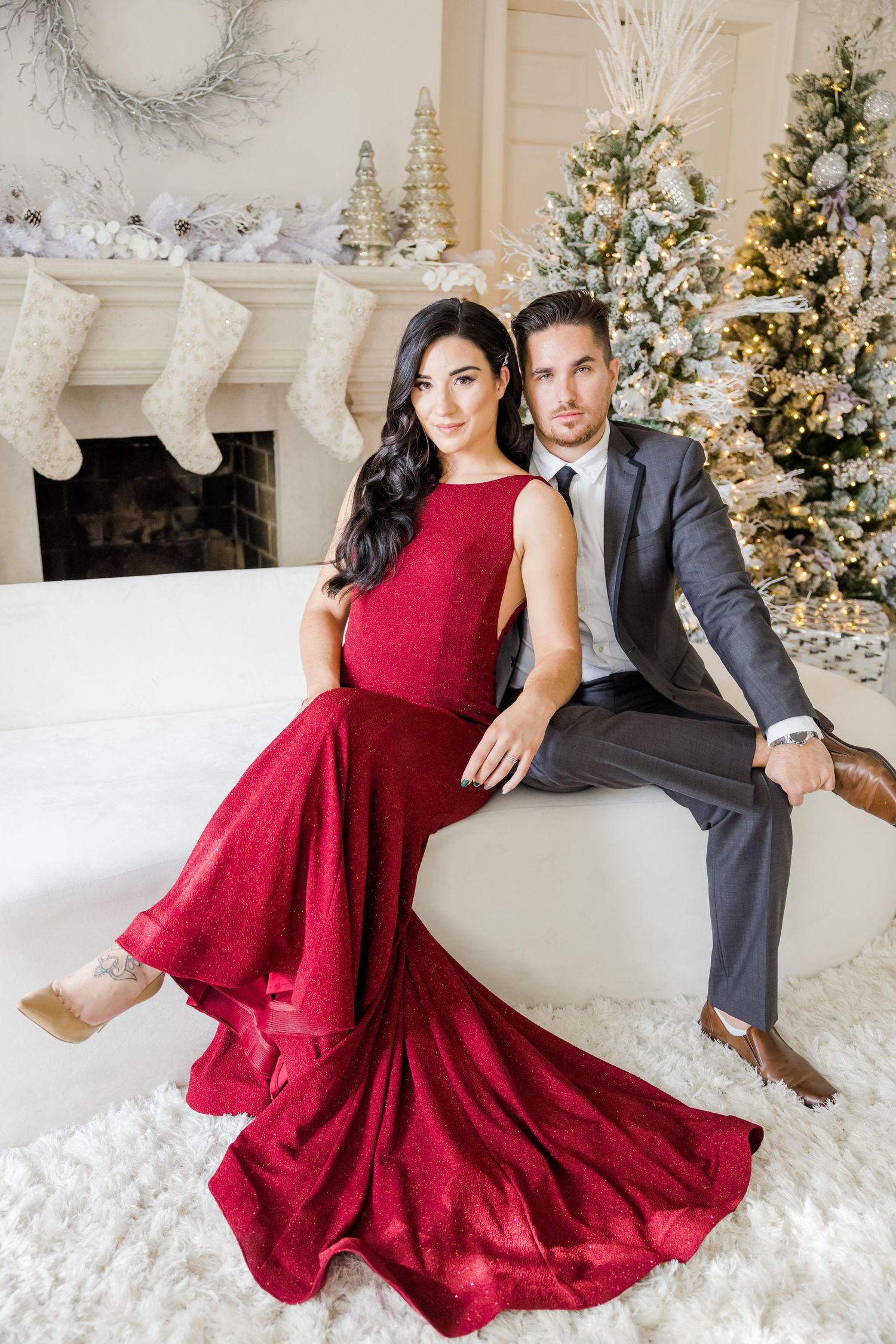 Romantic Christmas portrait with dramatic red gown and grey suit against decorated fireplace setting.