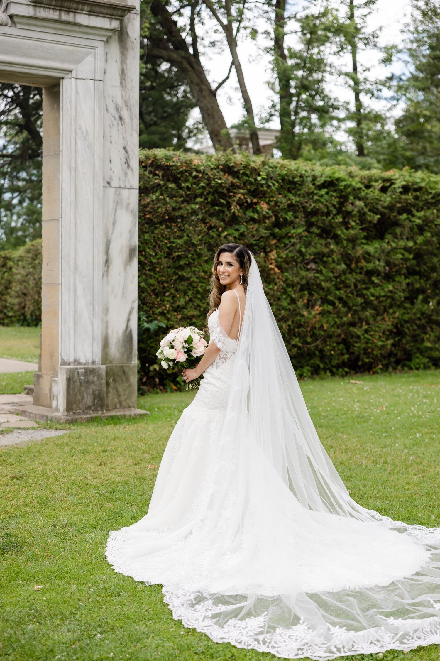 Dramatic cathedral length train and veil flow behind elegant wedding dress near classical stone archway.
