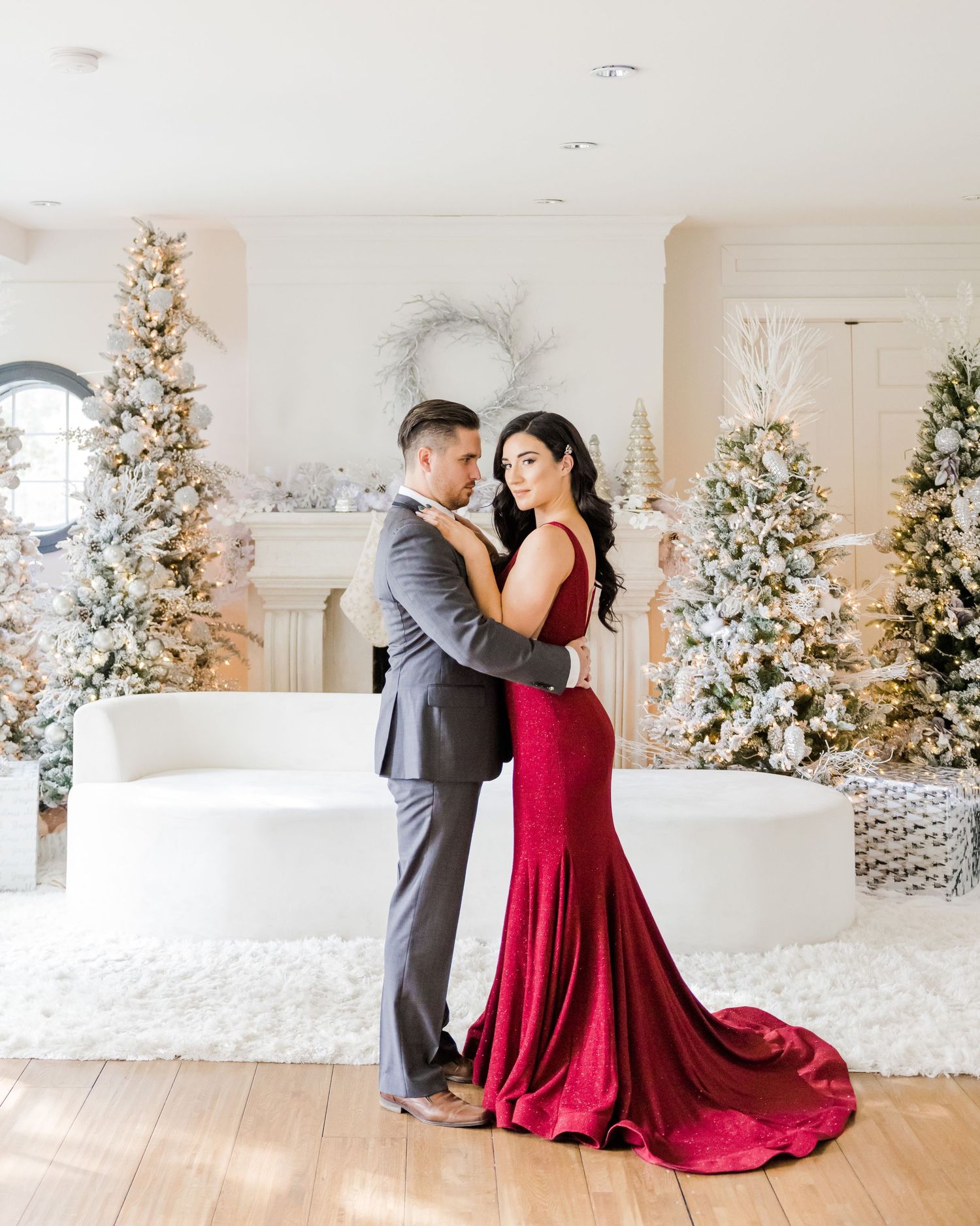 A romantic holiday photoshoot featuring a red mermaid dress and grey suit against snowy Christmas decor.