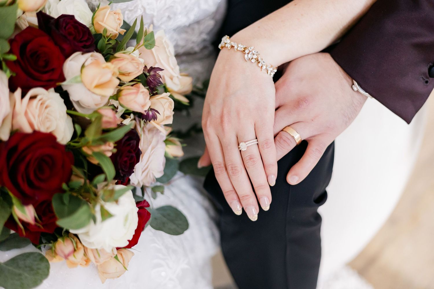 Close-up of wedding rings and hands with burgundy and blush floral bouquet.