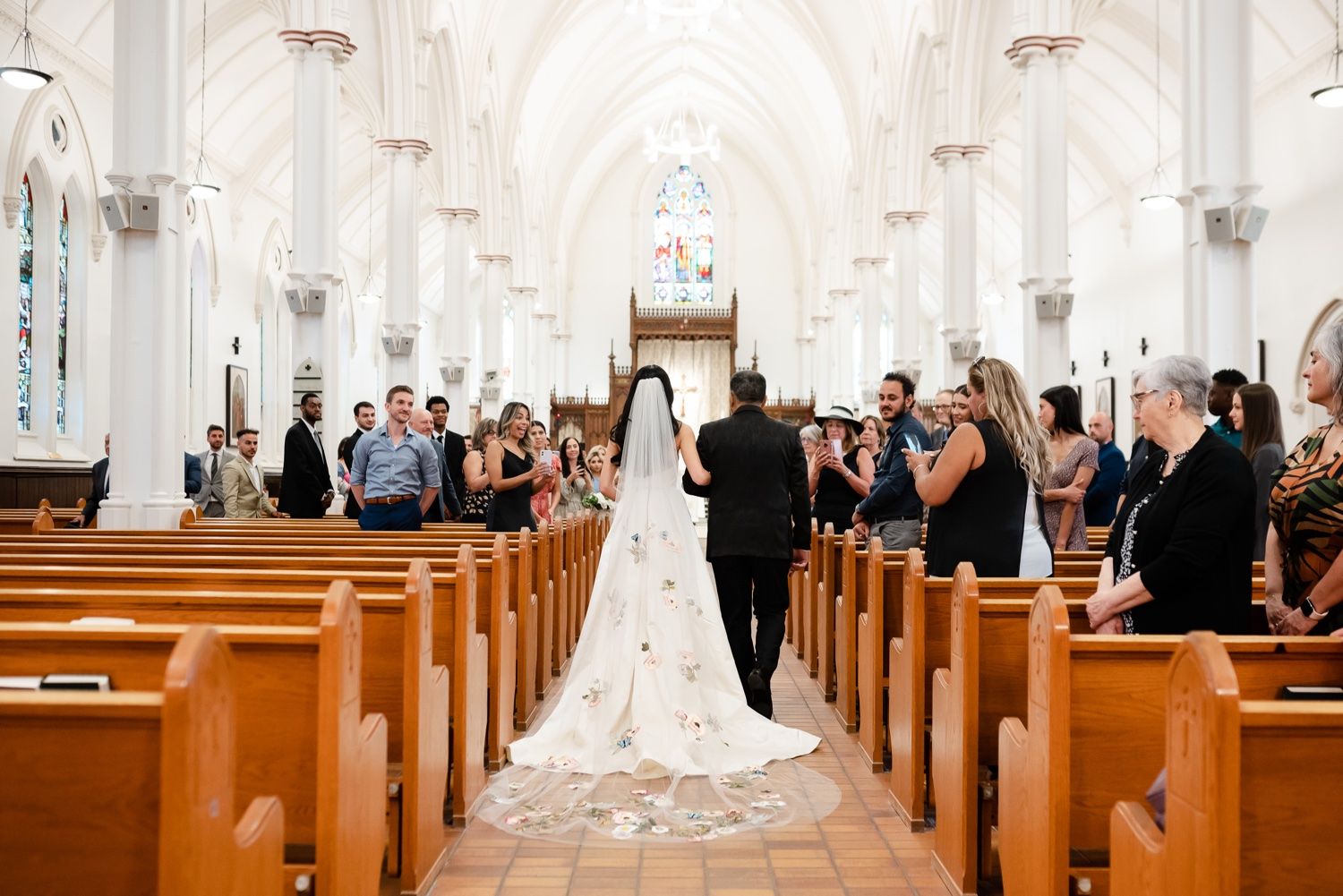 A wedding ceremony in a bright white cathedral church with guests standing in wooden pews along a long aisle.