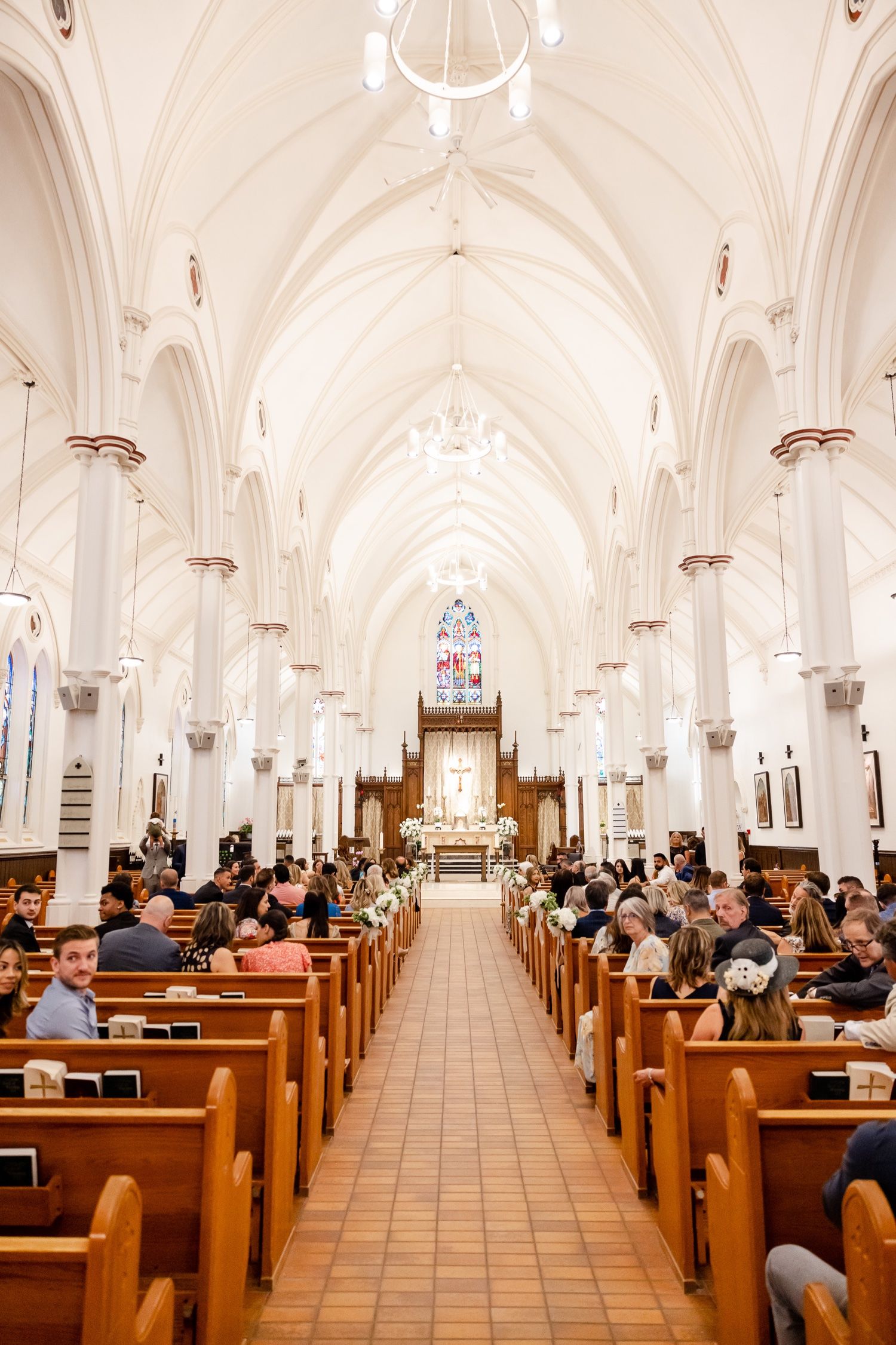 Interior view of a traditional church with white vaulted ceilings and wooden pews filled with wedding guests.