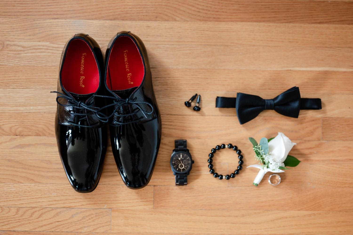 Wedding accessories including patent shoes, bow tie, watch, and boutonniere laid out on wooden floor.