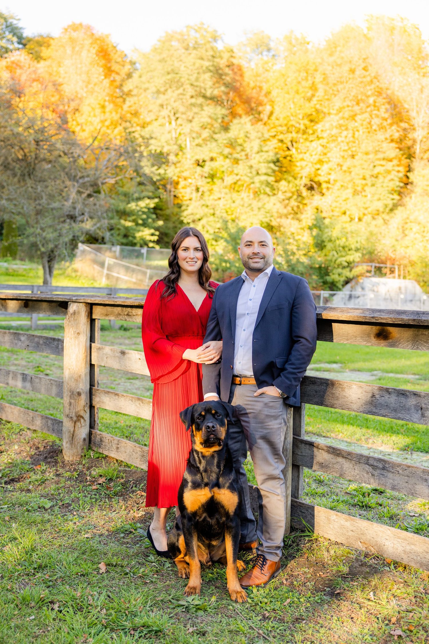 A couple in formal wear stands with their Rottweiler by a wooden fence during autumn.