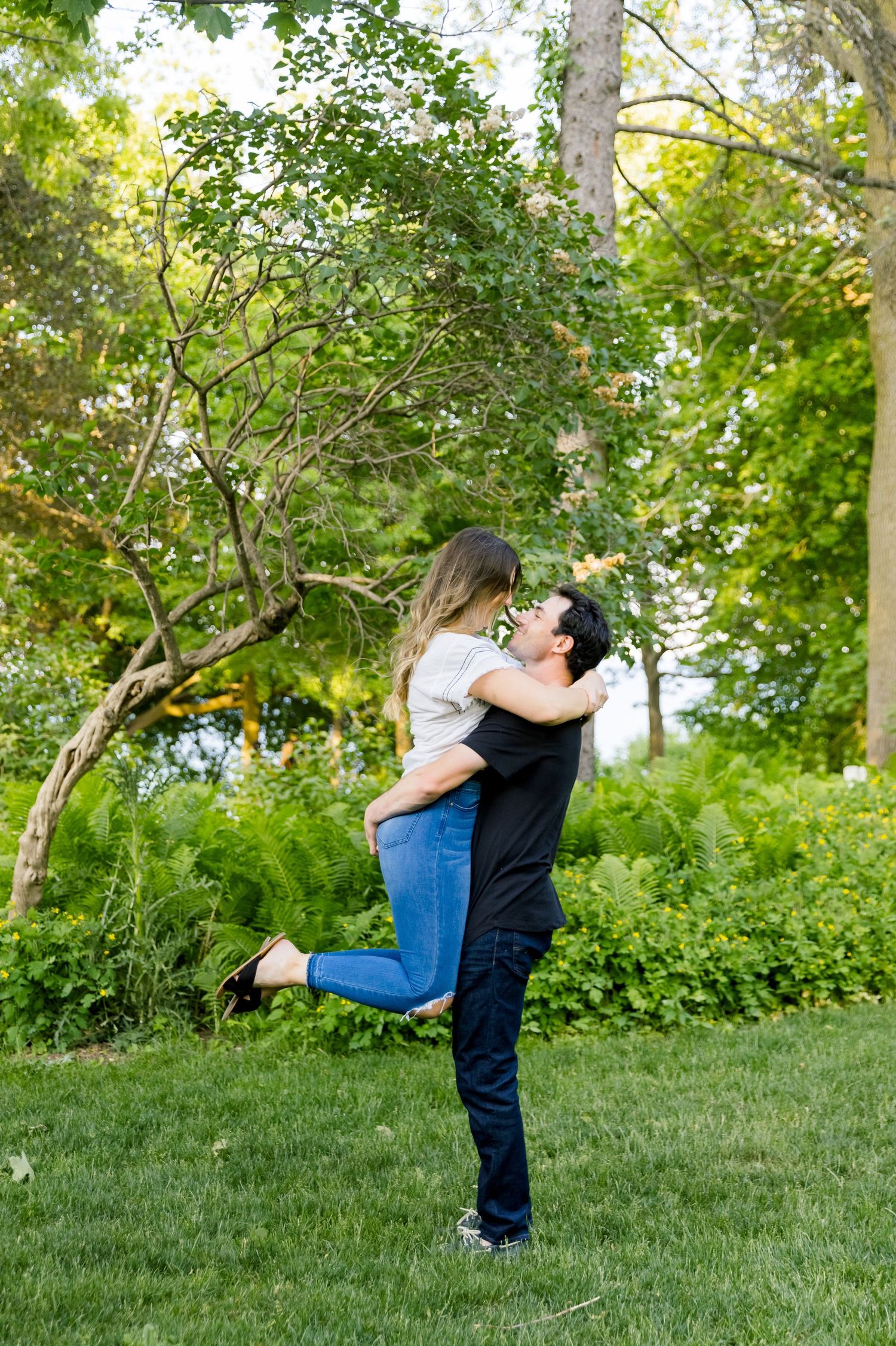 A couple embraces in a playful lift pose against a backdrop of lush green trees.
