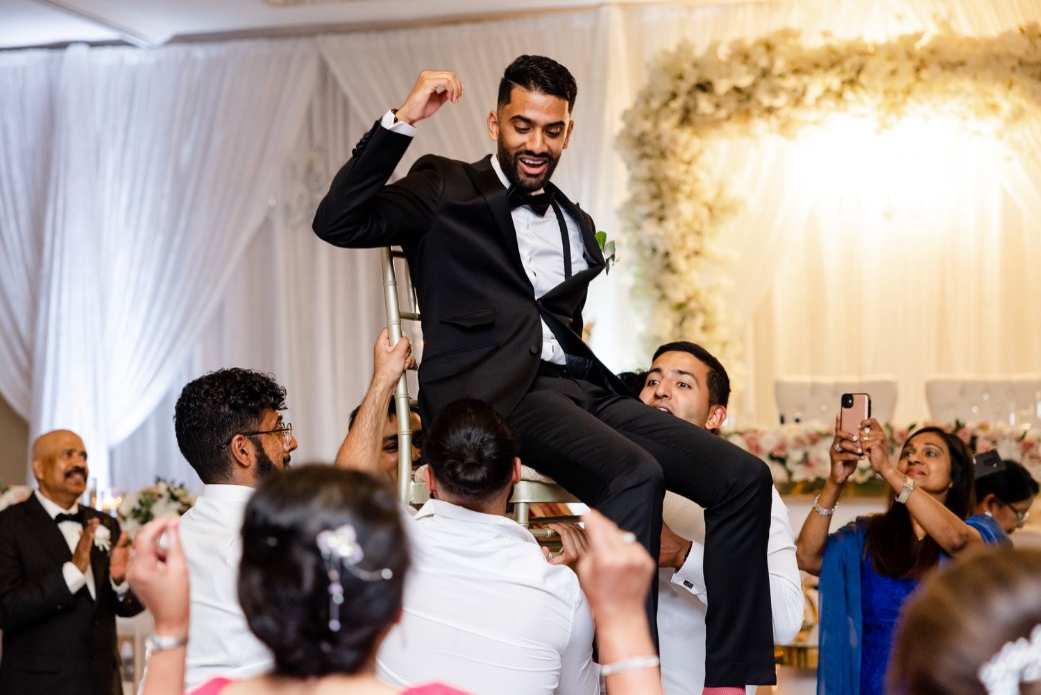 Person in black tuxedo being lifted on chairs during lively wedding celebration.