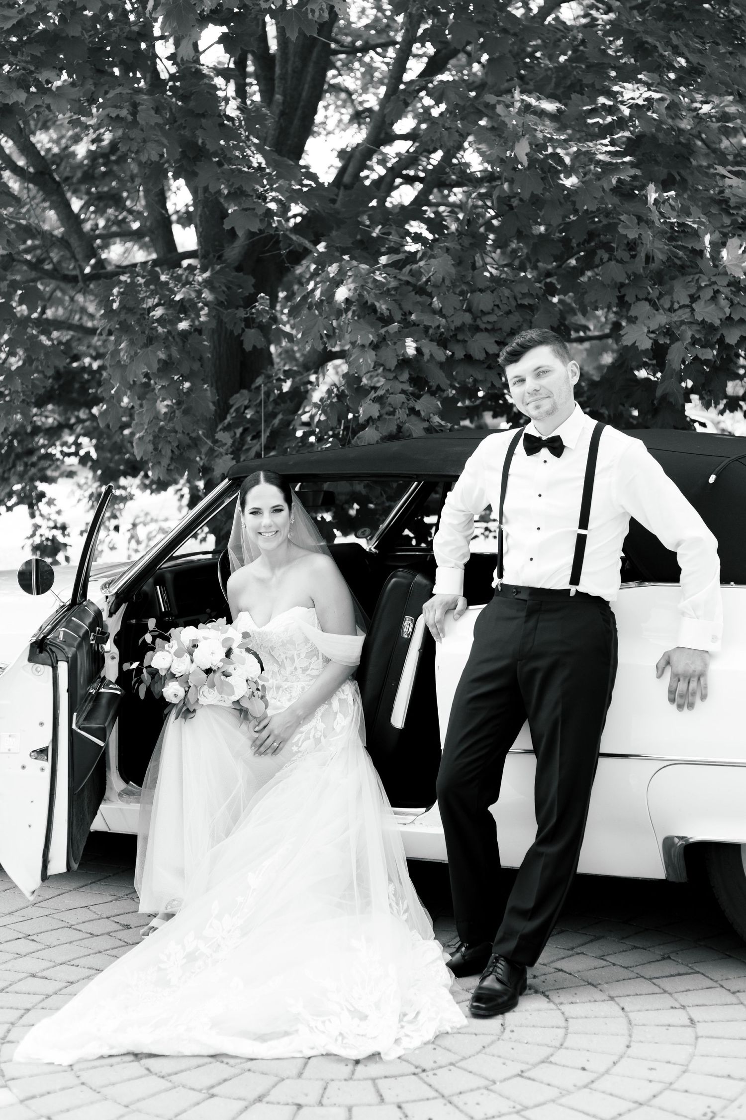 Classic wedding photo featuring a white classic car, with a trailing lace dress and formal attire against a leafy backdrop.