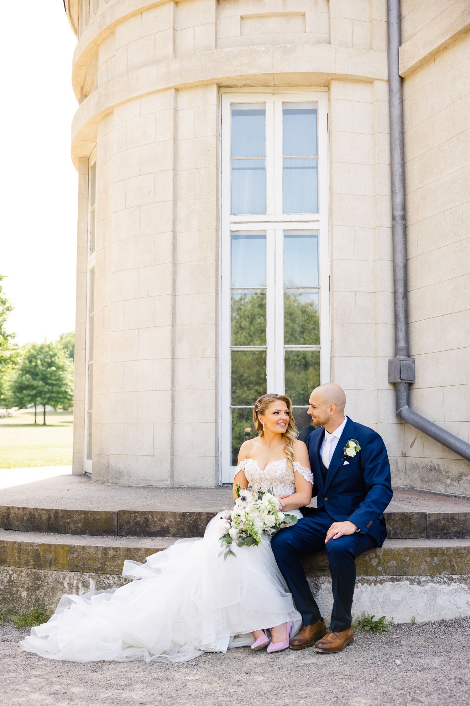 Newlyweds sit together on stone steps outside a grand building with a white dress and navy blue suit.