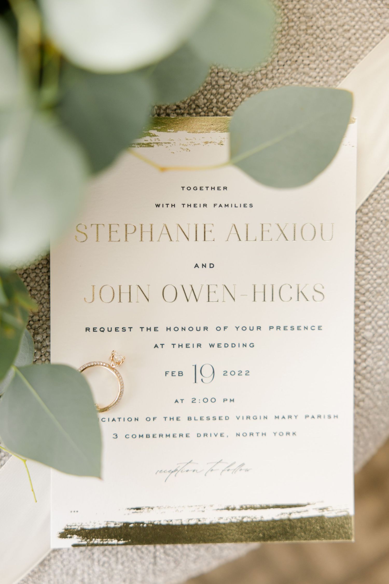 Modern wedding invitation with gold text and eucalyptus leaves on textured fabric.