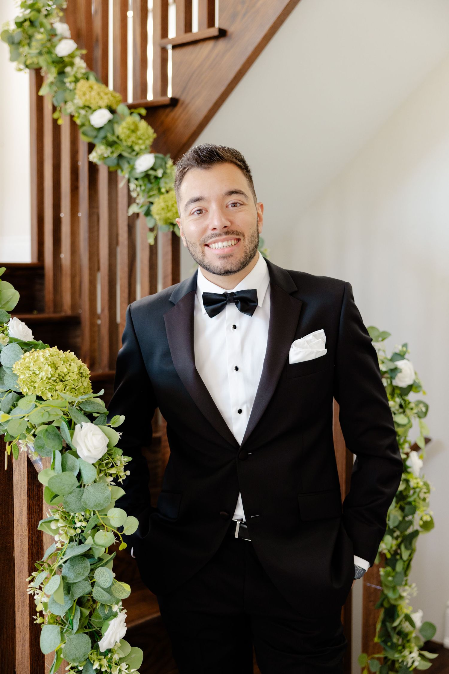 Groom in black tuxedo and bow tie poses by wooden staircase decorated with green floral garland.