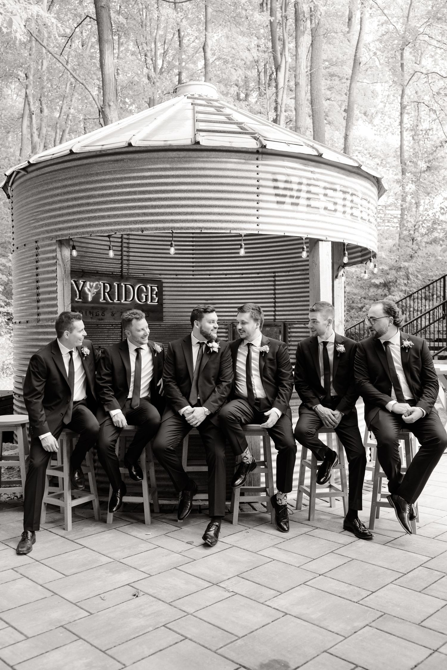Groomsmen in black suits sitting on wooden stools against metal silo bar structure in black and white.