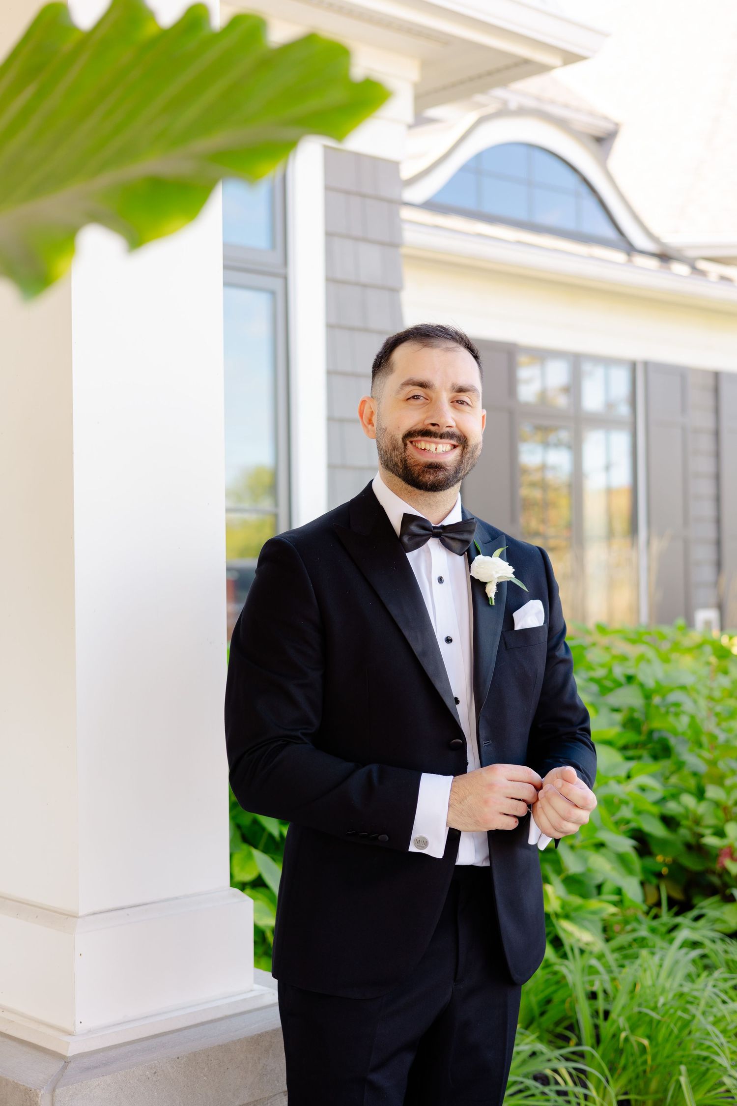 Groom in black tuxedo with white boutonniere smiling against architectural backdrop.
