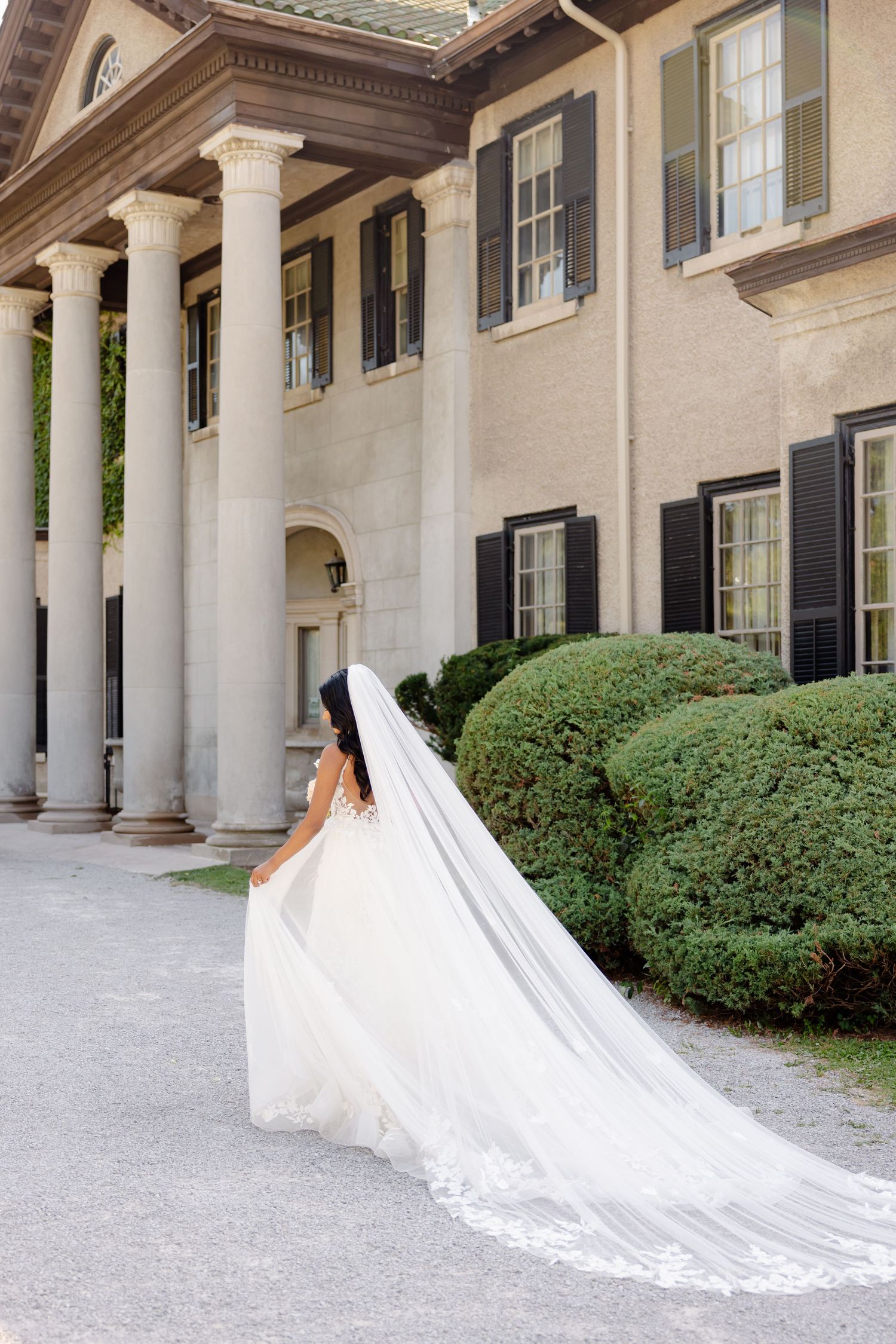 An elegant cathedral-length veil trails behind a white wedding gown against a colonial-style mansion backdrop.