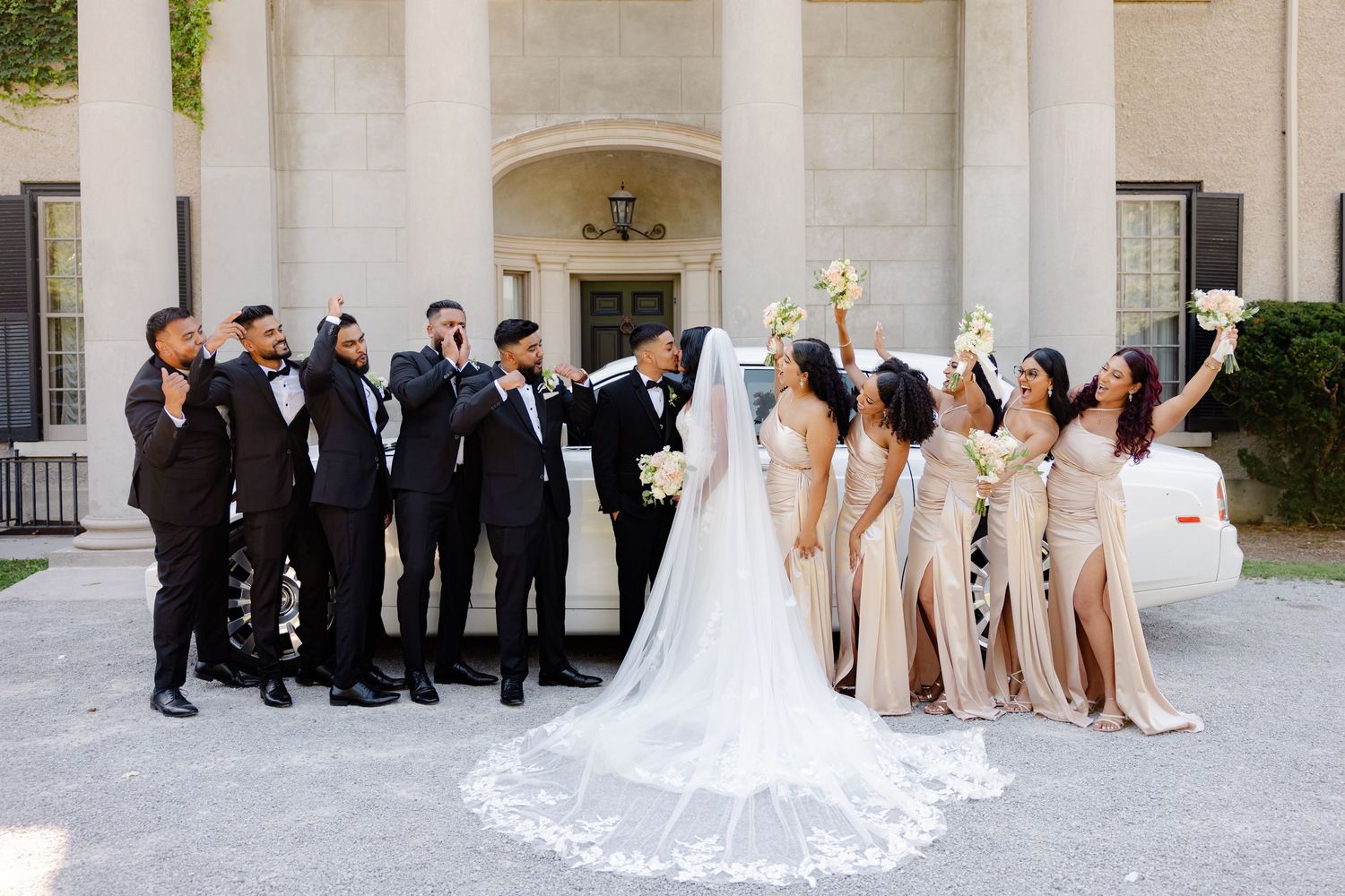 Celebratory wedding party photo with bridesmaids and groomsmen in front of elegant building.