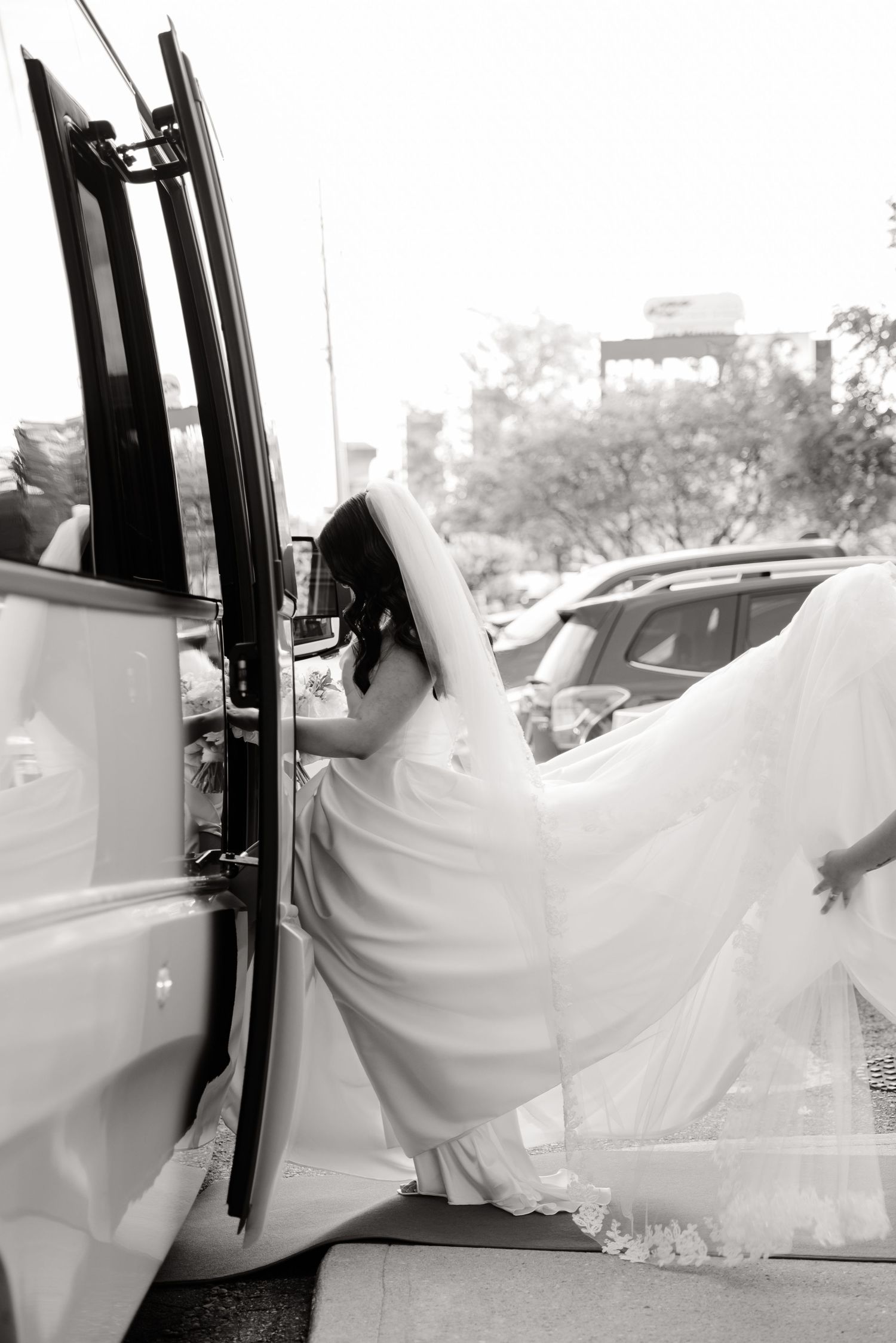 Black and white photo of a bride stepping into a vehicle in her wedding dress.