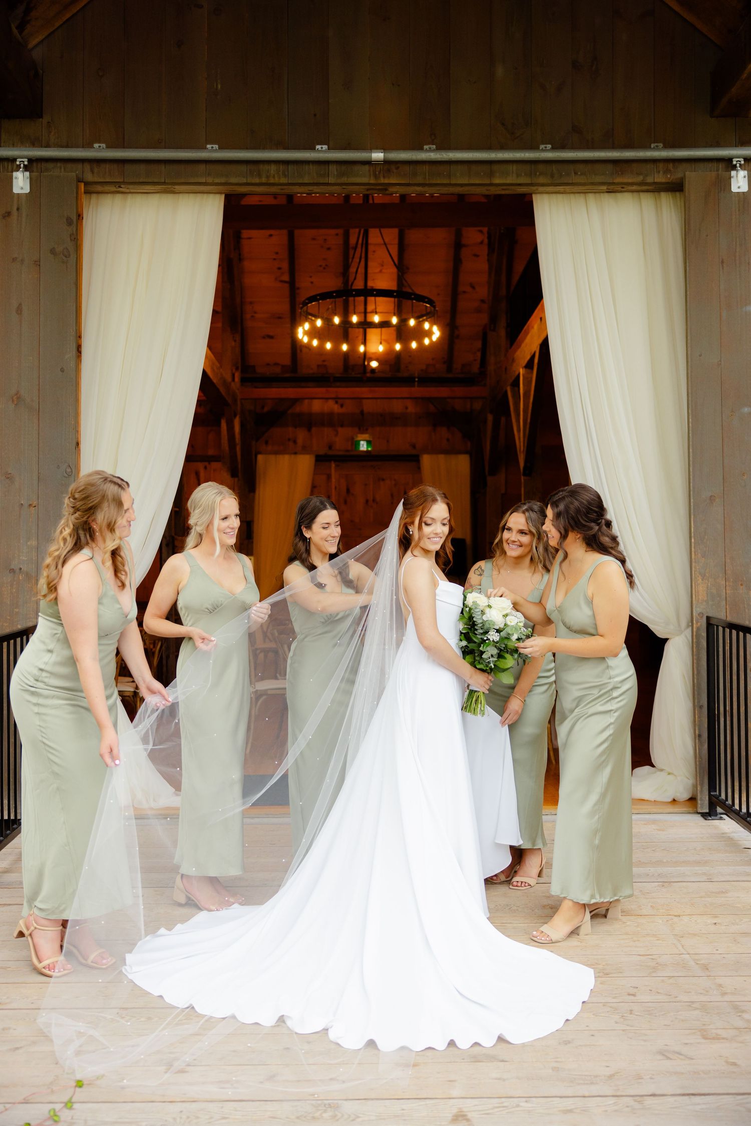 Bridesmaids in sage green dresses helping with bride's veil at barn venue entrance.