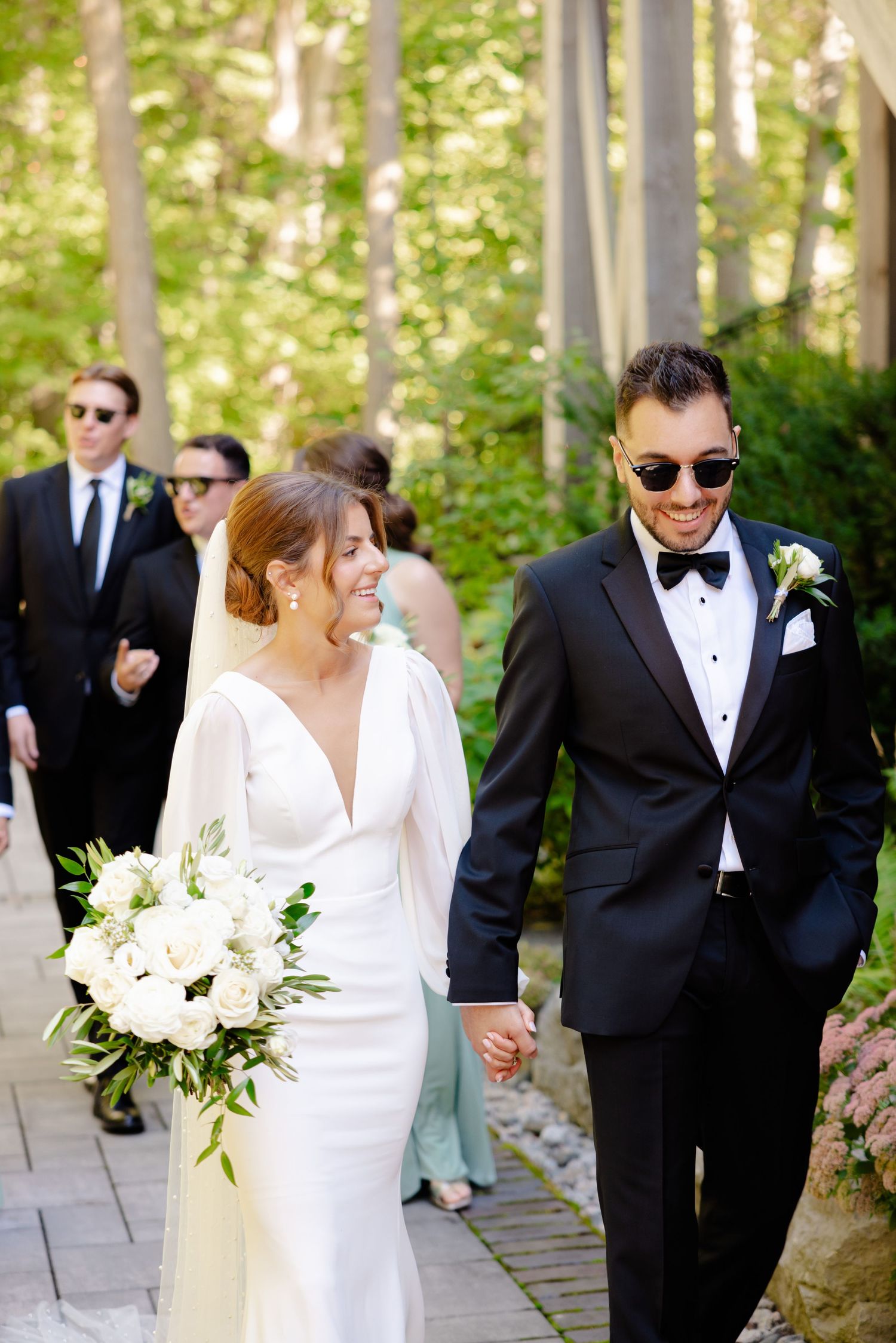 Newlyweds walking outdoors with wedding party, both wearing sunglasses and formal attire.