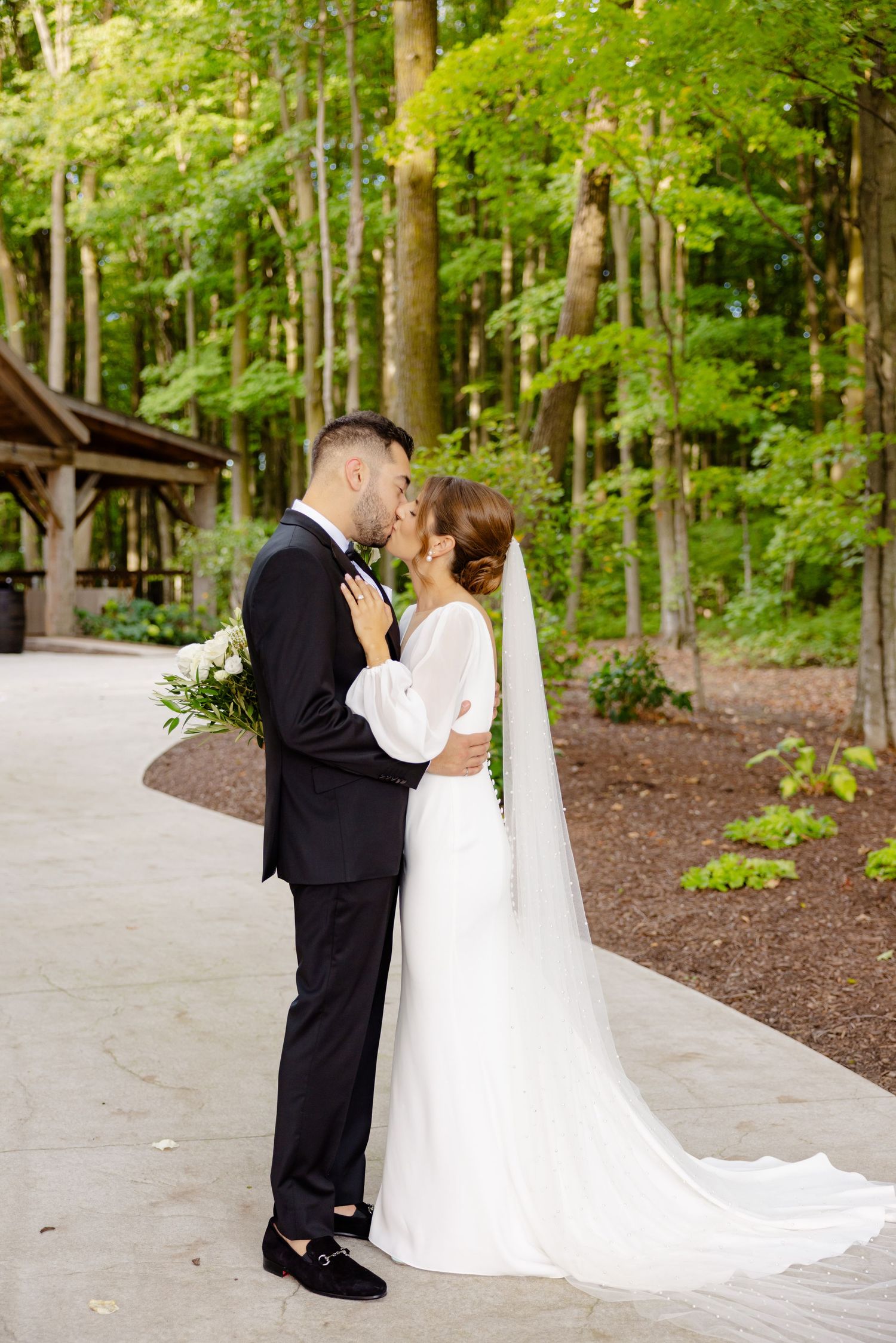 Newlyweds share a tender kiss on a concrete path surrounded by lush green forest.