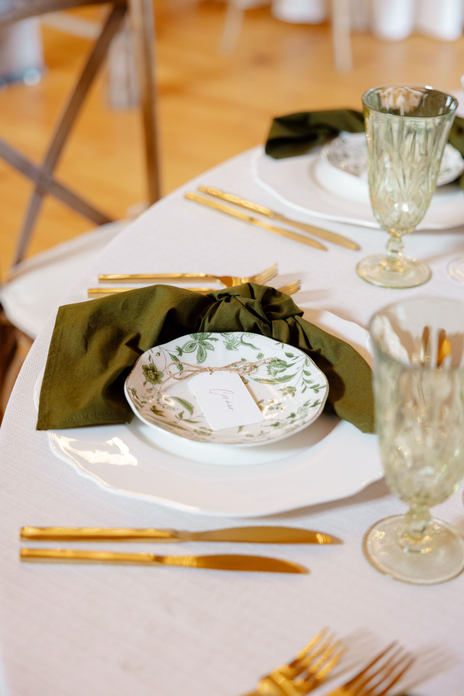 Wedding place setting with olive napkin, vintage plate, and gold cutlery on white tablecloth.