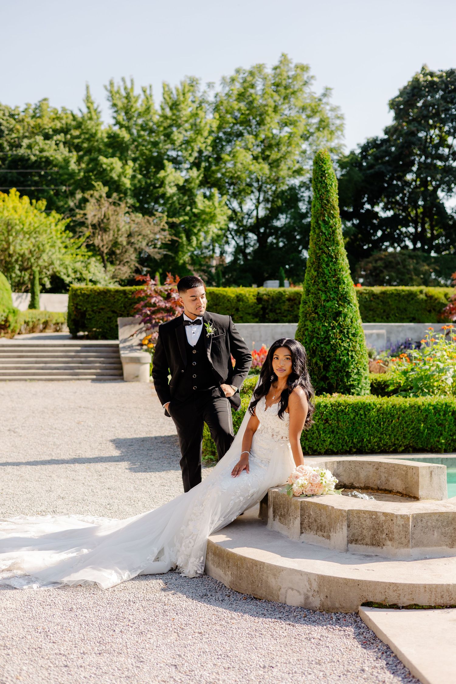Bride and groom pose on stone steps in an elegant garden setting with cypress trees.