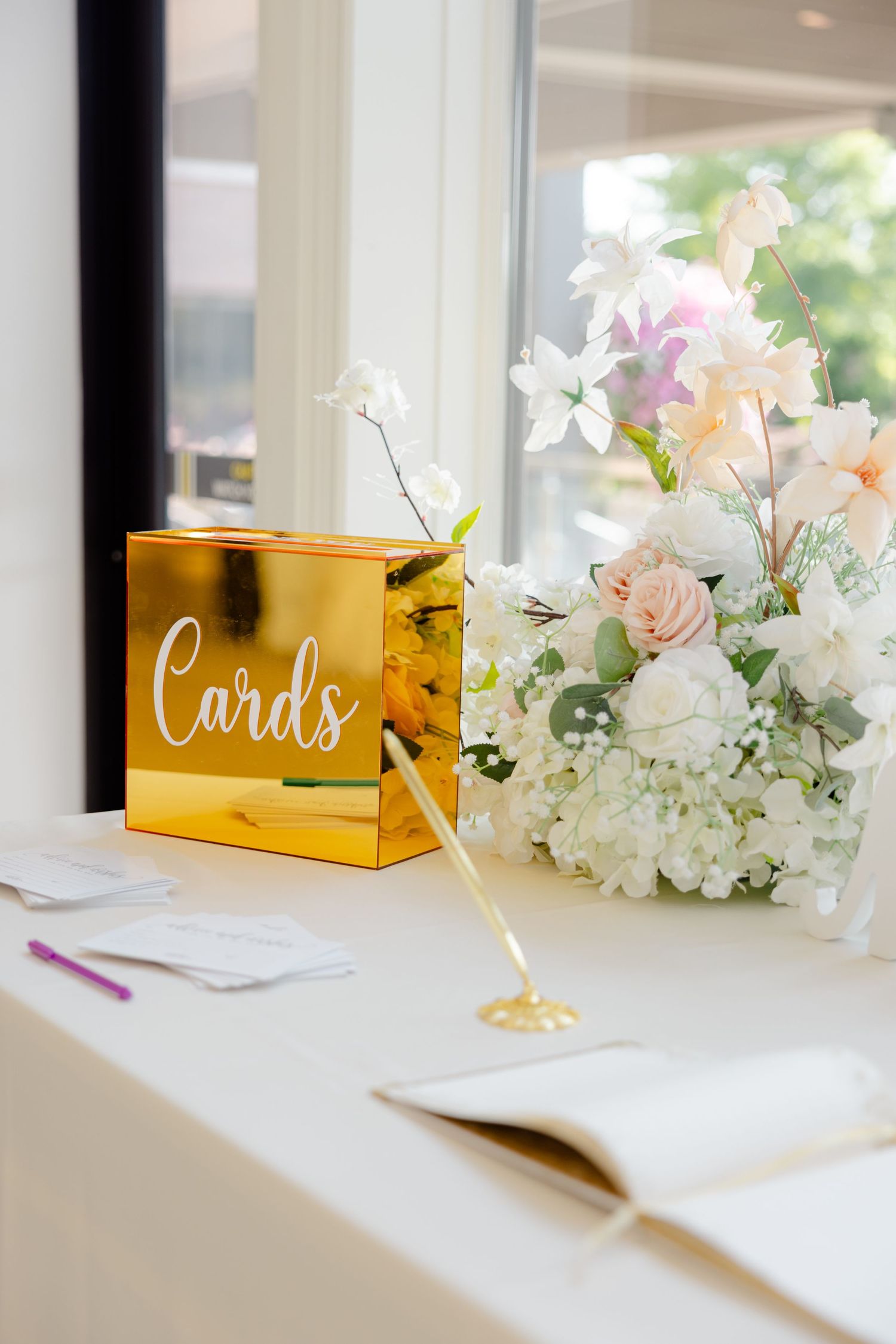 Elegant gold card box and light floral centerpiece arrangement on white table at wedding reception.