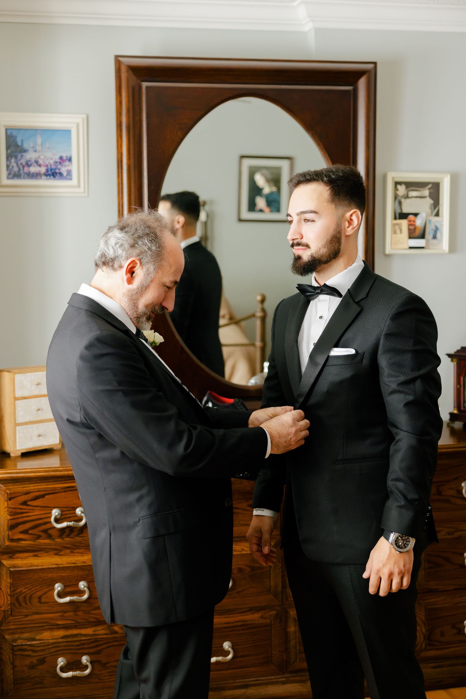 A touching moment of formal attire preparation shows adjusting of a bow tie against a wooden dresser backdrop.