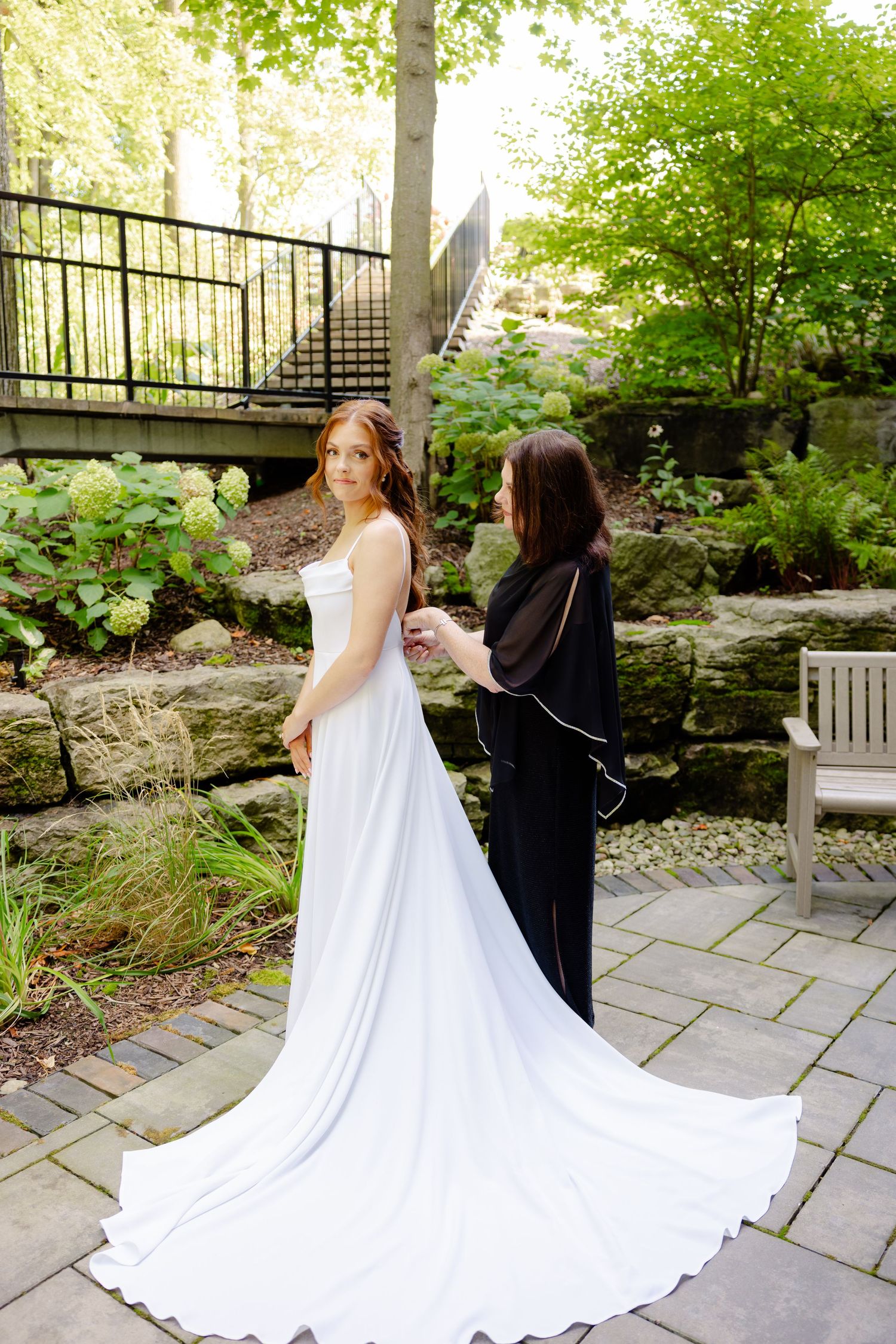 Elegant bride with flowing train poses in garden with assistant helping.
