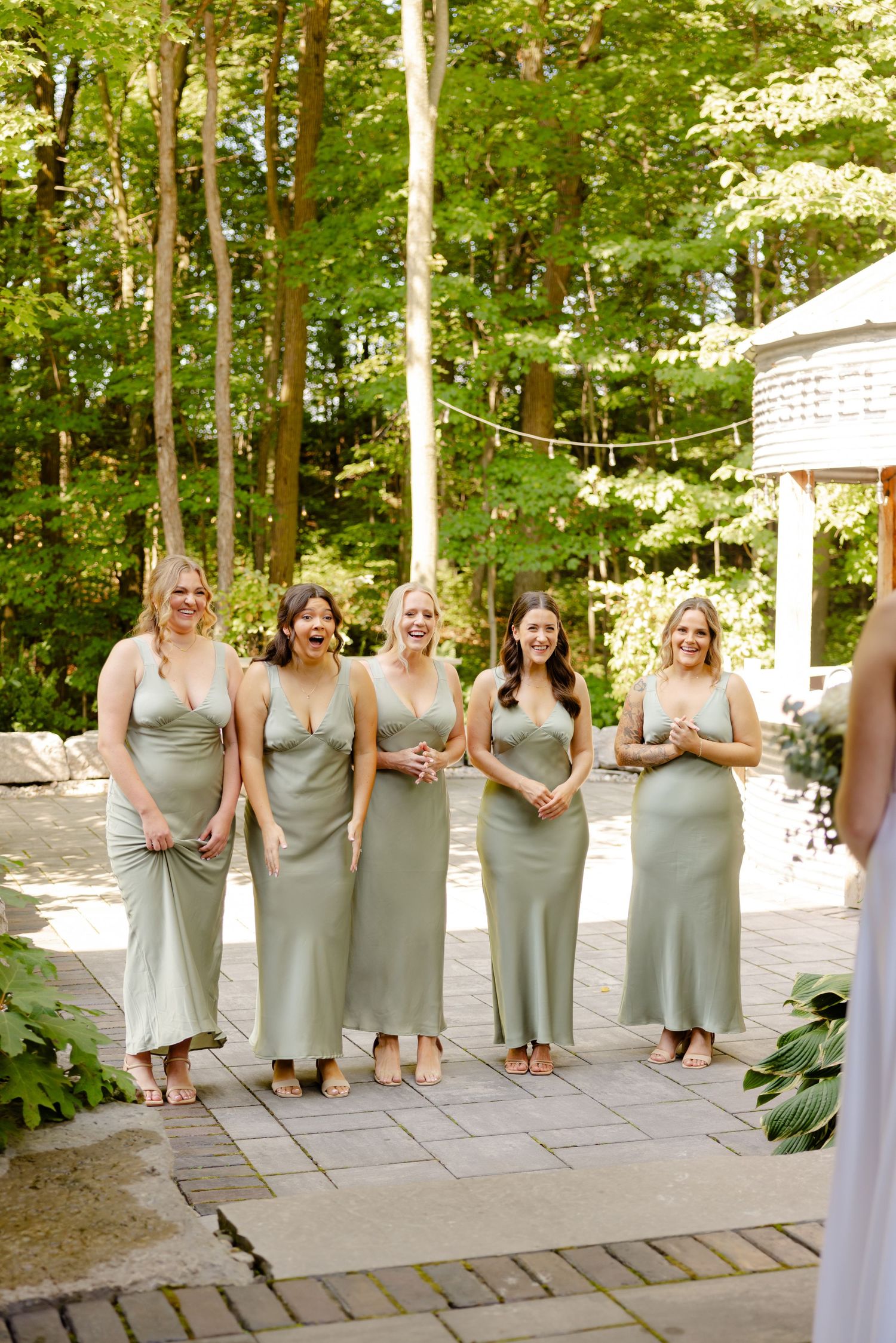 Bridesmaids in sage green dresses stand together on stone patio surrounded by lush garden foliage.