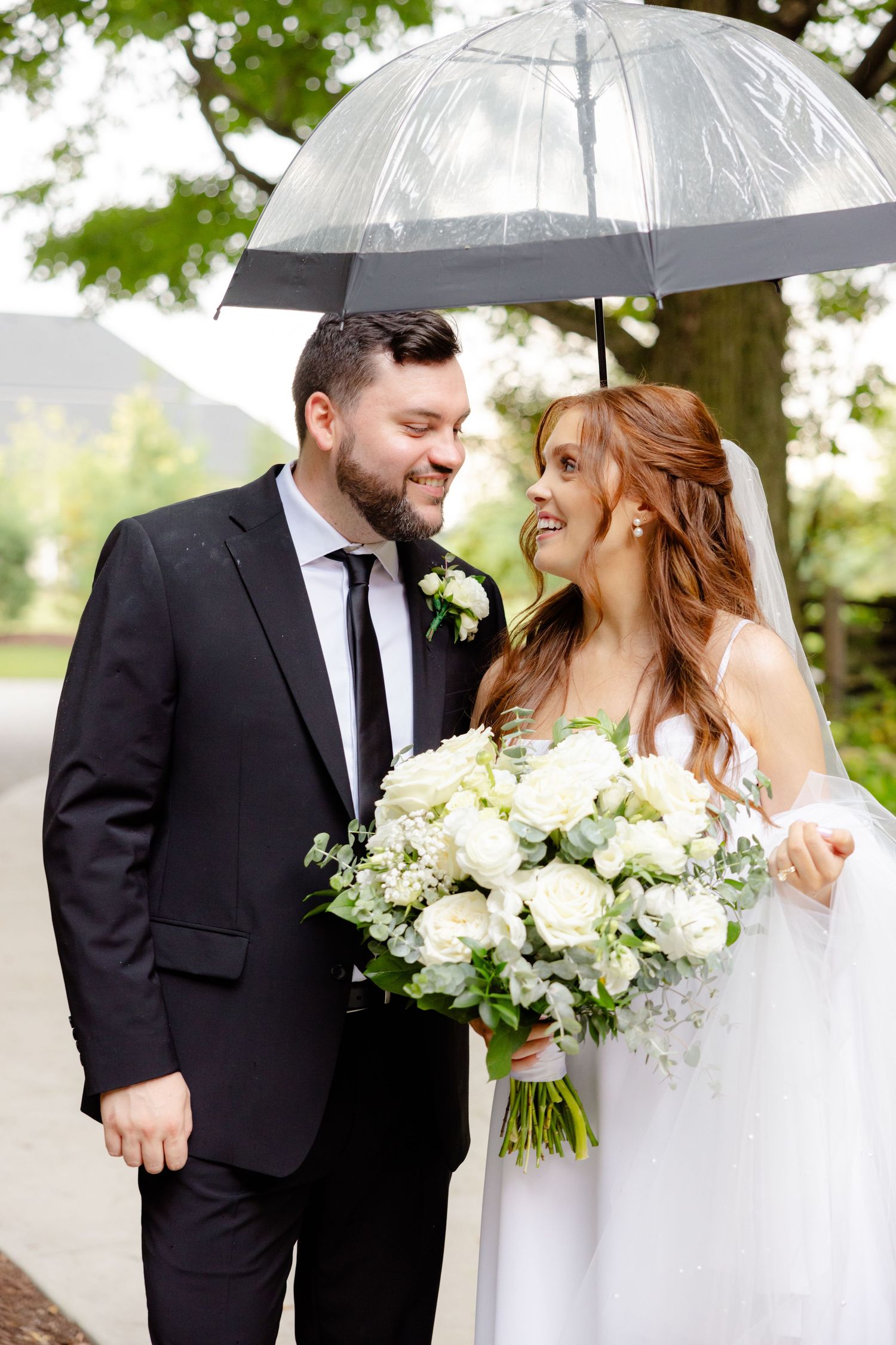 Happy couple shares intimate moment under clear umbrella while holding white floral bouquet.