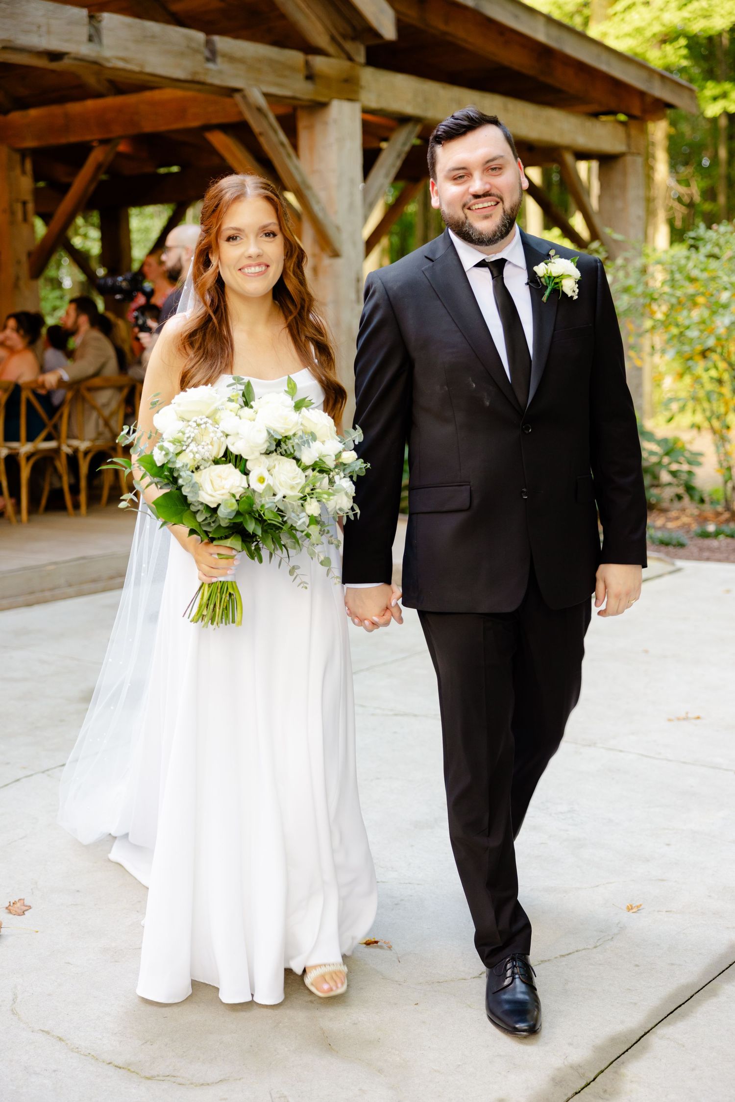 A wedding couple walks hand in hand along a concrete path with a rustic wooden structure in background.