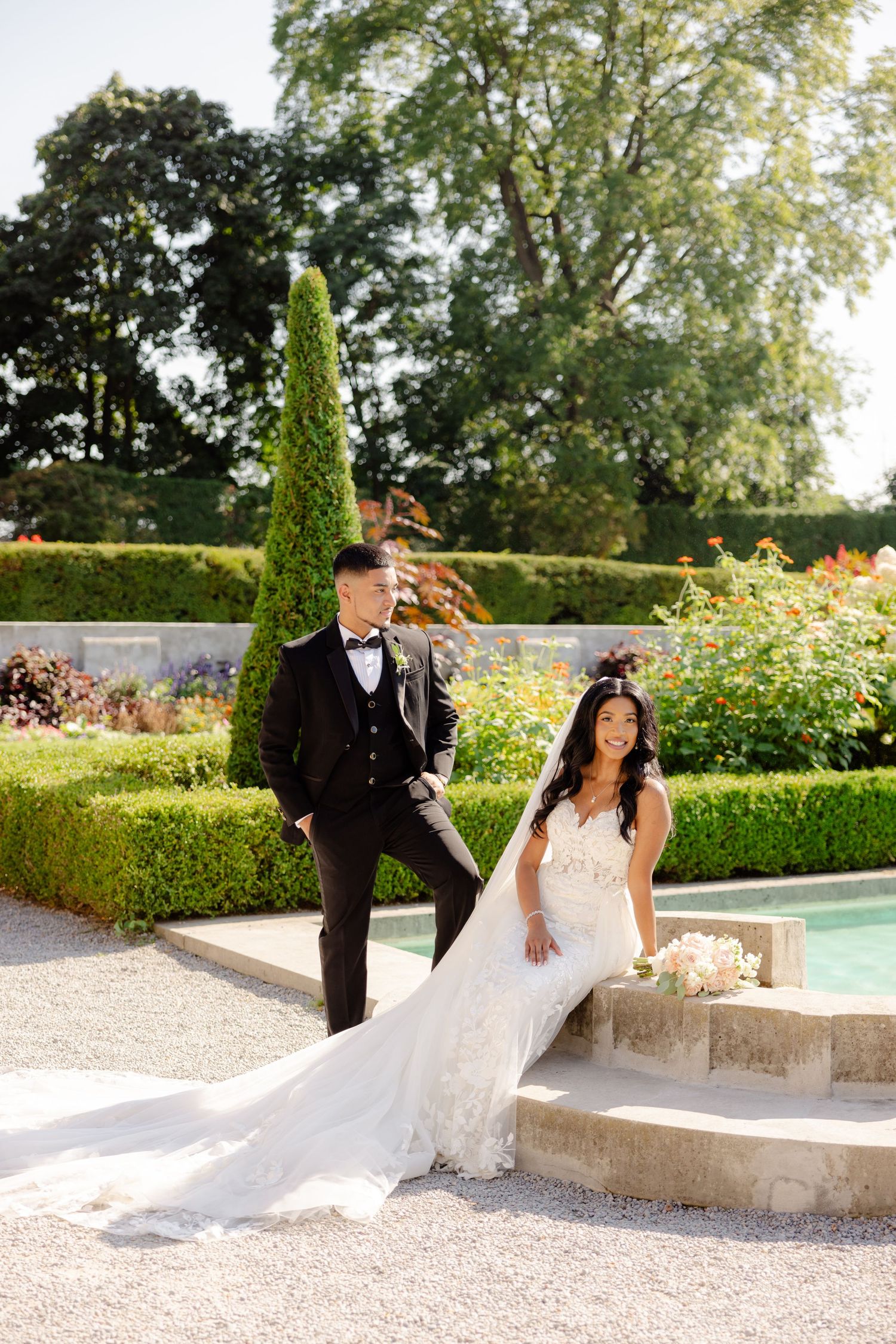 A couple in formal wedding attire poses beside an ornamental pool with sculpted cypress trees.