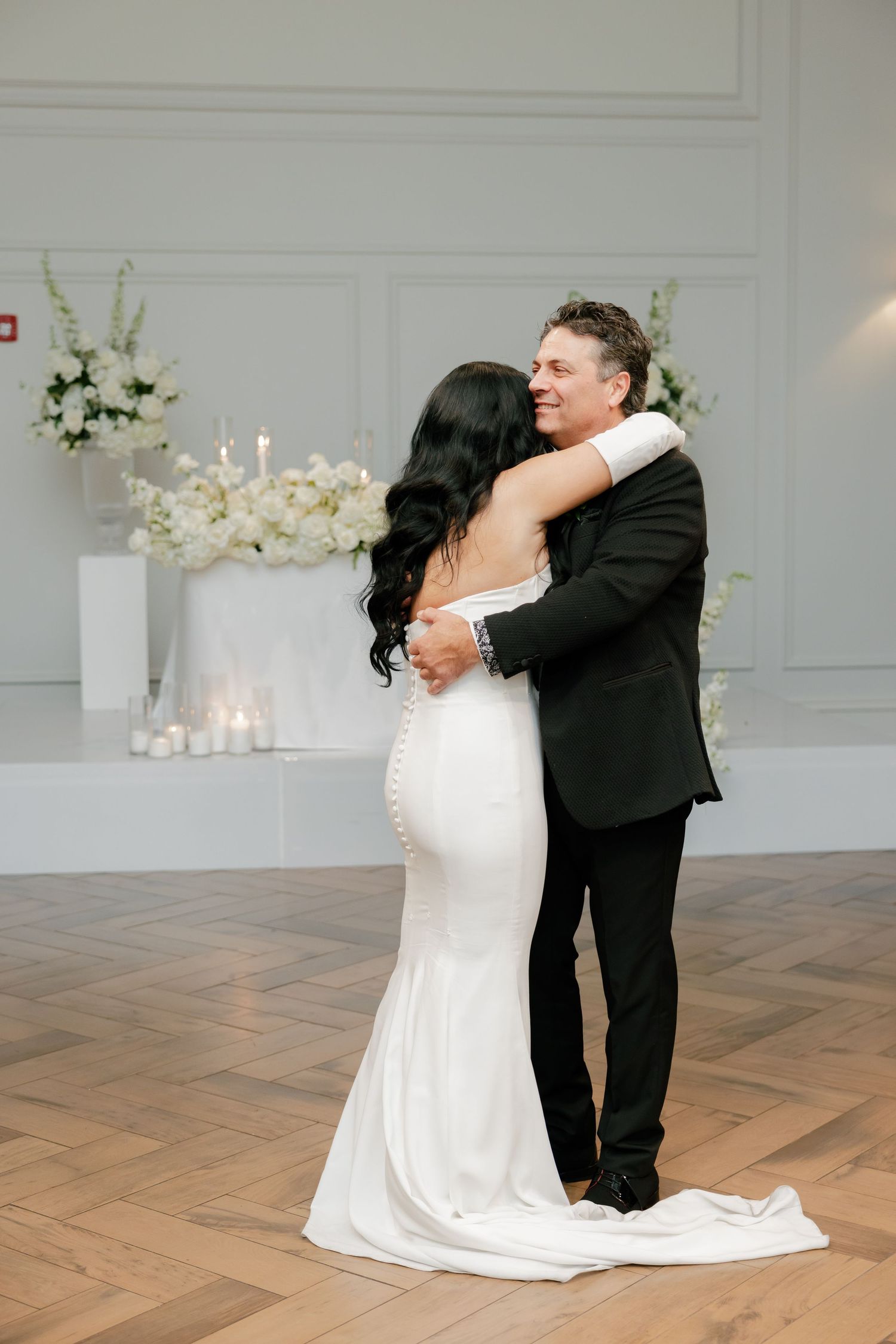 A bride in a fitted white gown and groom in black tuxedo share their first dance in an elegant white reception venue.