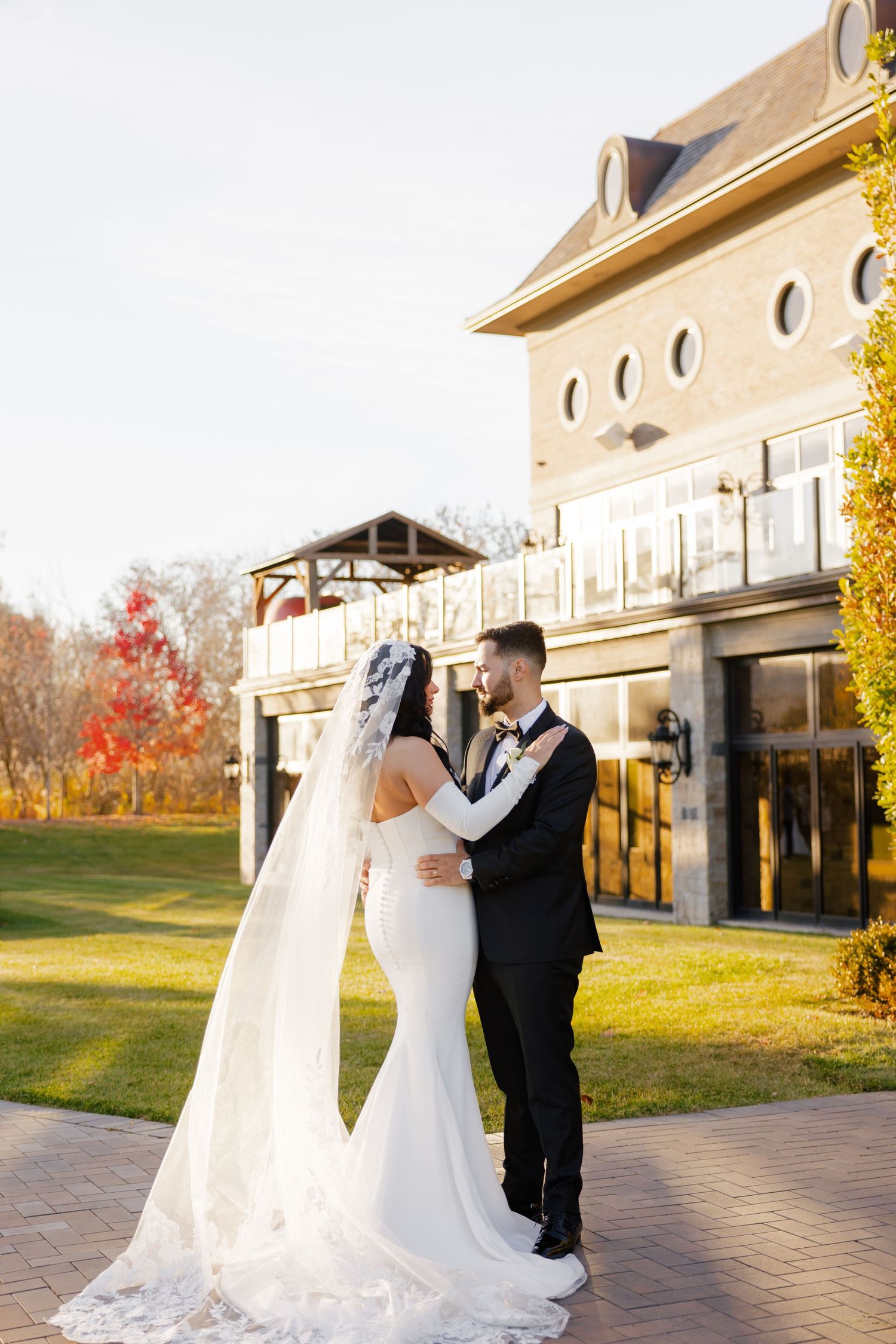 Wedding couple embraces outside stone building with circular windows and fall foliage.