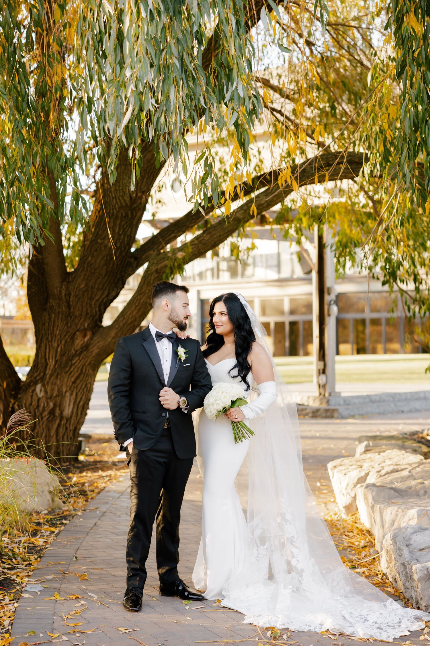 A romantic wedding photo of a couple standing beneath a weeping willow tree with autumn leaves.